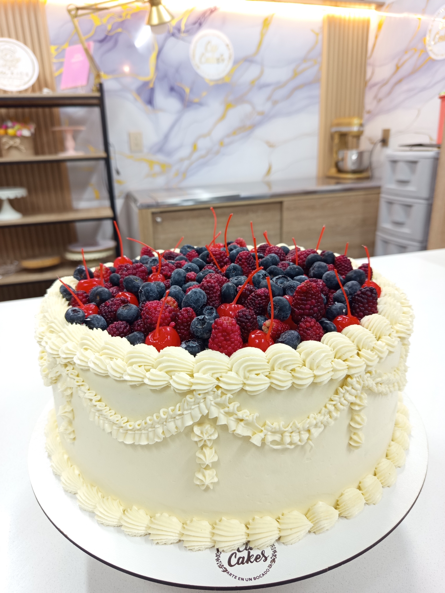 The image shows a white frosted cake topped with a generous amount of fresh berries, including raspberries and blueberries, and garnished with maraschino cherries. The cake is decorated with intricate buttercream piping along the sides, forming a scalloped border at the base and a decorative swag pattern higher up. A circular logo with the text "Cakes Arte en un Bocado" is visible on the cake stand. In the background, there is shelving with cake stands and decorative items, a marble-patterned wall with gold accents, and kitchen appliances. The scene appears to be set in a bakery or a dessert shop, potentially in Girón, Colombia, given the text on the logo.