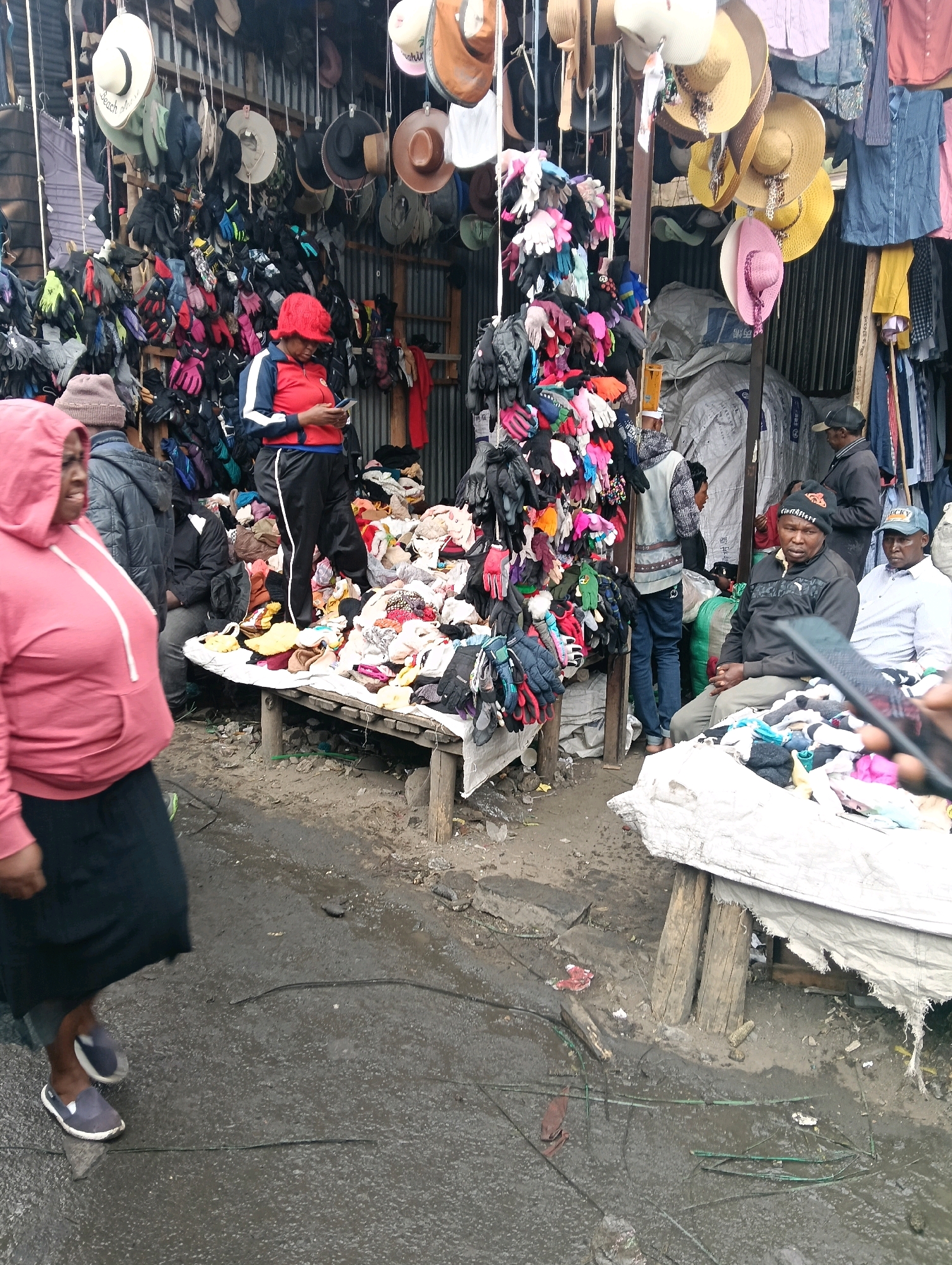 This image captures a bustling outdoor market scene in Nairobi, Kenya, likely a roadside stall. The foreground shows a woman in a pink hooded jacket and black skirt walking, her feet clad in blue and white slip-on shoes. She is moving from left to right, partially out of focus. Behind her, a man in a grey jacket and brown beanie is visible.
The midground is dominated by a market stall selling a wide array of items, primarily hats and gloves, hanging from racks and displayed on tables. A woman in a red jacket and black pants stands behind a table laden with various garments and accessories, looking down at her phone. Several other individuals are present in the background, some appearing to be vendors or customers. The stall is set up under a makeshift shelter, with corrugated metal and fabric sheeting providing cover.
The environment appears to be a somewhat disorganized and potentially wet area, with exposed ground and what looks like puddles or wet patches on the ground. The overall impression is of a vibrant, informal market economy typical of many urban areas in Nairobi. The scene is crowded with goods, suggesting a high volume of merchandise being offered for sale. The lighting suggests it is daytime.