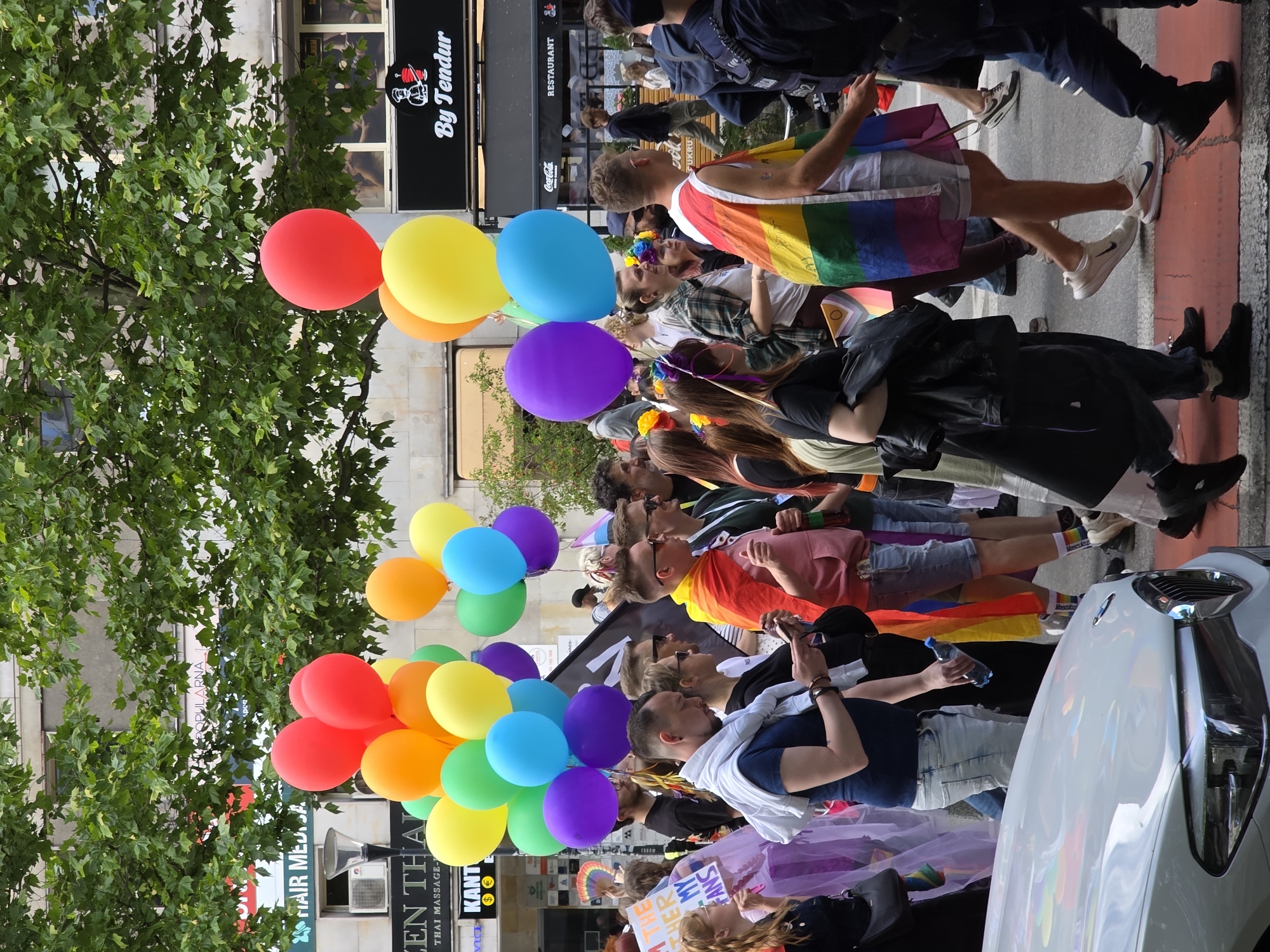 This is a medium shot of a pride parade taking place on a city street, likely Warsaw, Poland, given the presence of "By Tendur" signage which is a restaurant chain located in Warsaw. The scene is bustling with people walking and carrying rainbow flags and balloons. In the foreground, a white car is partially visible. The participants are diverse, with many wearing colorful clothing and accessories. Some individuals are holding up rainbow flags, symbolizing LGBTQ+ pride. The background shows buildings and trees lining the street, indicating an urban environment. The overall atmosphere appears festive and celebratory.