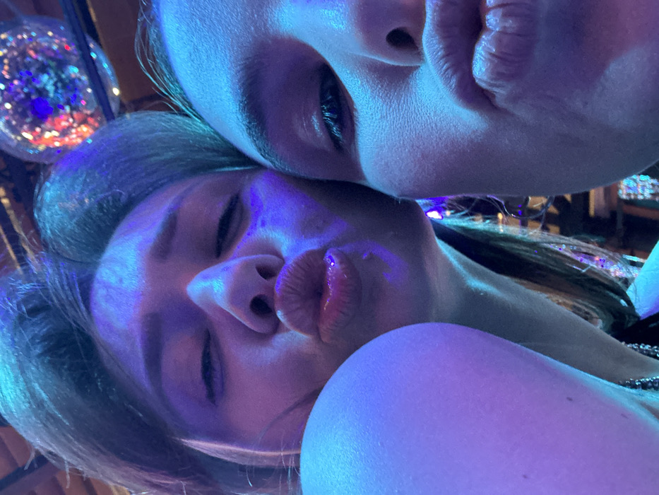 The image displays a close-up selfie of two females in an indoor setting. Both subjects are positioned in close proximity, facing the camera with their lips pursed in a pouting expression. The individual on the left has closed eyes, while the individual on the right has their right eye partially visible. The scene is illuminated by artificial blue and purple lighting, creating distinct color casts on their faces and hair. In the upper right background, a multi-faceted disco ball is visible, reflecting various colored lights, indicating a dynamic, social environment such as a nightclub or bar. No specific landmarks or identifying features are present within the image to determine a precise location in Moscow, Russia.