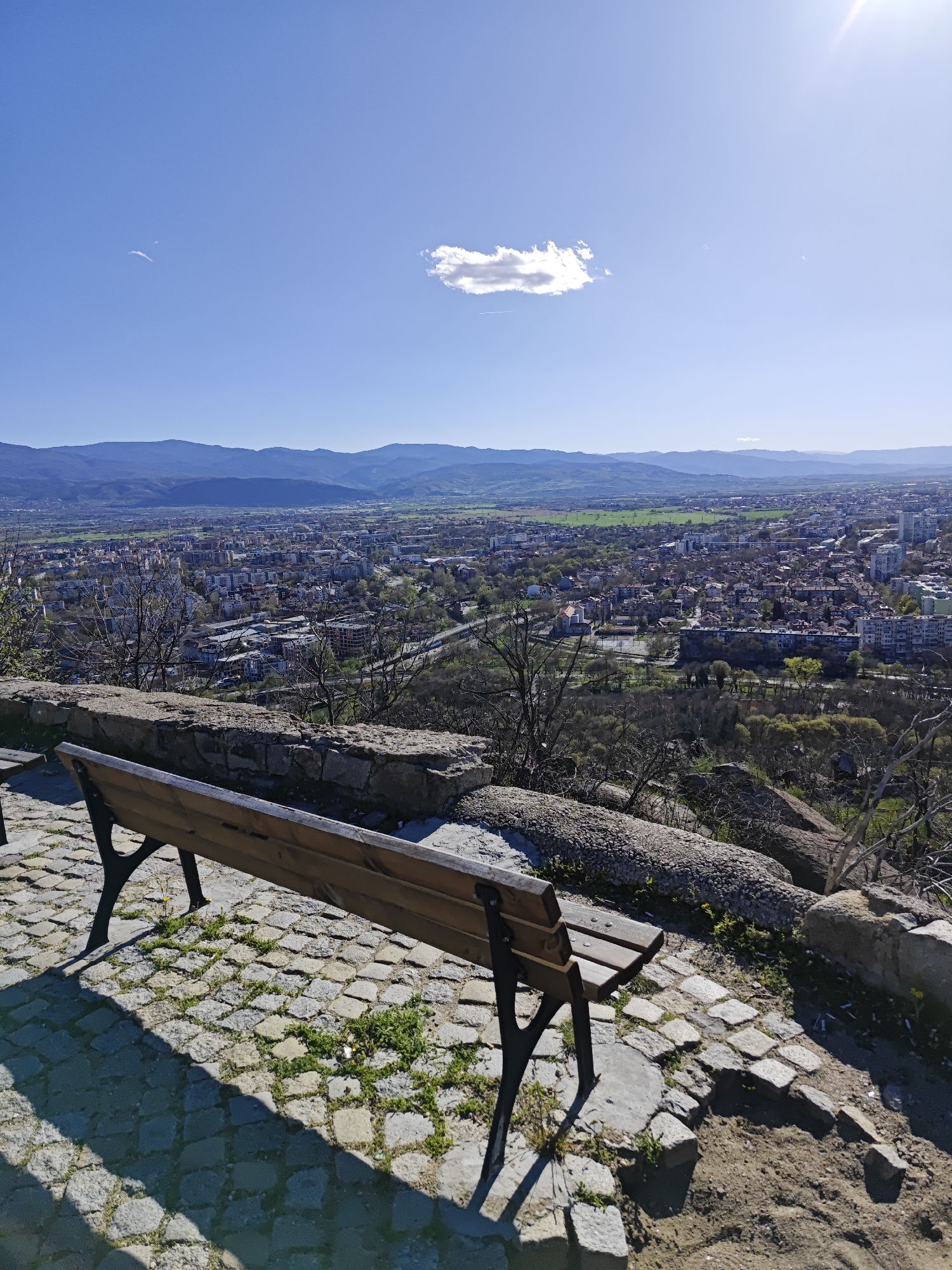 A daytime, long-shot, eye-level photograph captures a scenic view from Nebet Tepe in Plovdiv, Bulgaria. A weathered wooden bench with black metal legs is positioned on a cobblestone pathway in the foreground. The background reveals an expansive cityscape of Plovdiv nestled in a valley, with a backdrop of rolling hills and mountains under a clear blue sky. A single cumulus cloud floats in the center of the sky, with the sun shining brightly near the upper right corner. Vegetation, including patches of green grass, sprouts between the cobblestones around the bench. Bare tree branches are visible in the middle ground, interspersed with stone remnants of ancient walls. The photograph's lighting casts shadows from the bench onto the cobblestone. No people are visible.