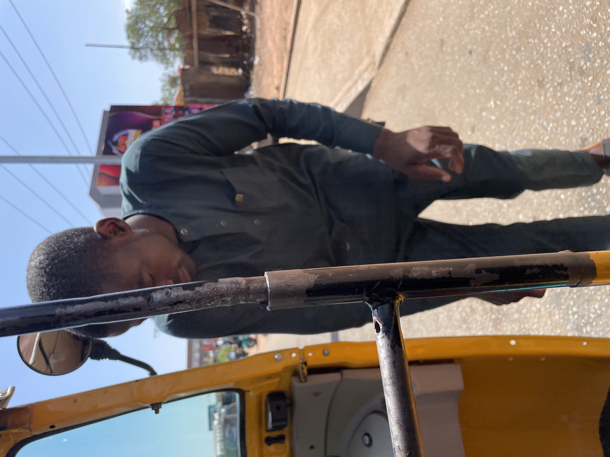 A man in Jos, Nigeria, rests his head on the safety bar of a yellow tricycle, likely used for transportation. He wears a long-sleeved, dark green uniform-style shirt and dark trousers. The vehicle is parked on a paved surface with buildings visible in the background. Power lines are overhead. A billboard is partially visible behind him. The sky is bright.