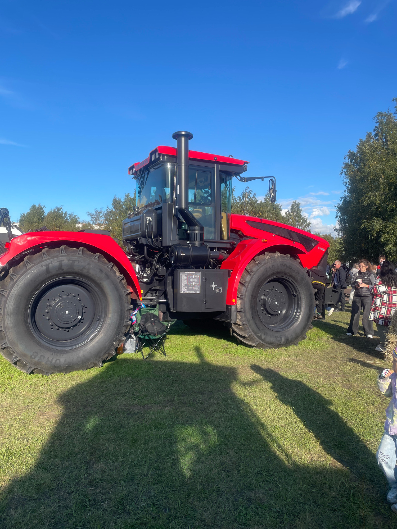 The image shows a large red tractor parked on a grassy field. The tractor has massive tires and a tall exhaust pipe. The cabin is enclosed with windows. Several people are standing in the background, some appearing to be onlookers or attendees at an event. The sky is blue with a few wispy clouds. Trees are visible in the background, suggesting an outdoor setting. The scene is brightly lit by sunlight, casting long shadows on the grass. A green folding chair and a black bag are near the tractor's front wheel. The ground is covered in green grass. In the far right, a child in a light purple jacket and blue jeans with "Hello Kitty" embroidered on them is visible. The presence of spectators and the tractor displayed outdoors suggest this could be an agricultural exhibition, a fair, or a demonstration event in an open area within or on the outskirts of Saint Petersburg, Russia, possibly during a pleasant season like late summer or early autumn given the foliage and clear sky.