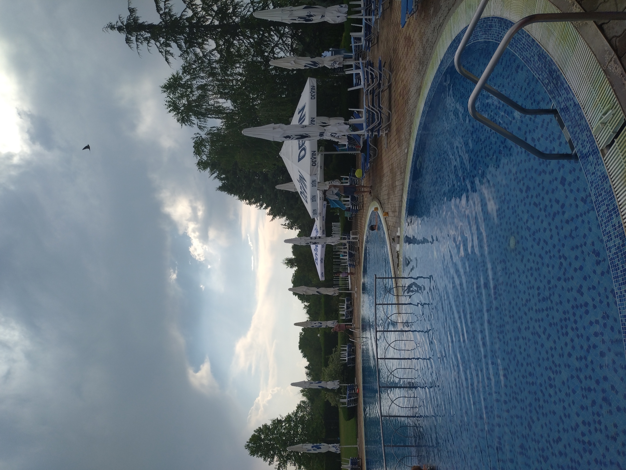 The image shows an outdoor swimming pool area at a resort in Yagoda, Bulgaria. The pool is filled with blue water and has a tiled bottom. A metal railing surrounds part of the pool. Several lounge chairs and large umbrellas are set up on the paved area around the pool. The umbrellas are white and feature the "Devin" logo. In the background, there are trees and lush greenery. The sky is cloudy, with patches of sunlight breaking through. A bird is visible flying in the sky. There are a few people present; one person is standing near the pool, and another appears to be in the water.
