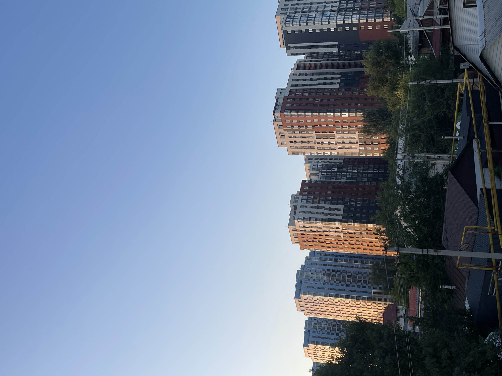 The image captures a cityscape with multiple residential buildings of varying heights and architectural styles under a clear blue sky. The buildings are a mix of modern high-rises, some with reddish-brown facades and balconies, and others with light blue and grey exteriors. Trees and greenery are visible in the foreground, interspersed with lower-rise structures and utility poles, suggesting a residential area within a city. The lighting indicates it is likely morning or late afternoon, with the sun casting warm tones on some of the buildings. Based on the context of Almaty, Kazakhstan, this scene could represent a view of the newer residential developments on the outskirts or within certain districts of the city.