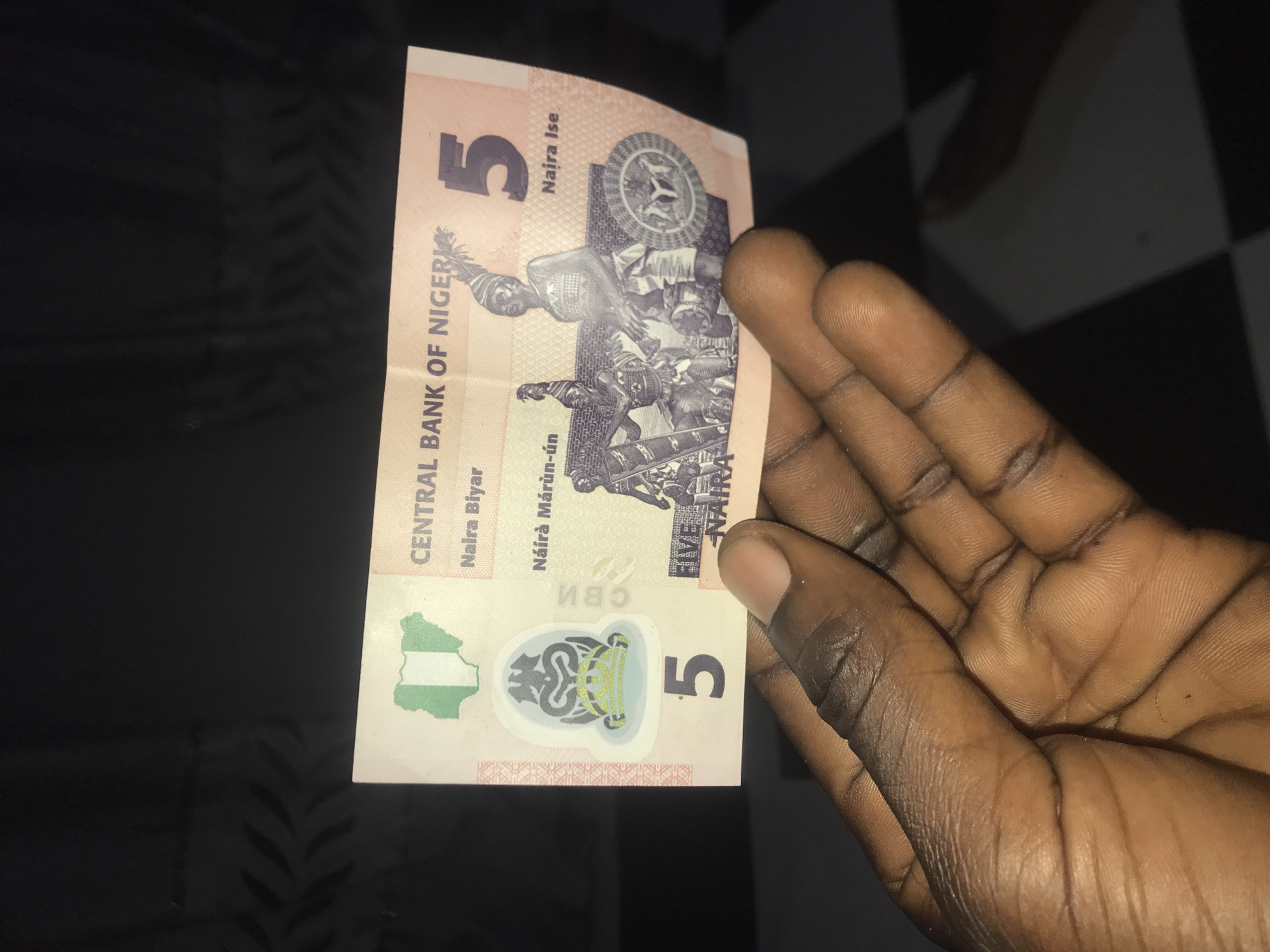 A person's right hand with dark skin tone is captured in a close-up, holding a single 5 Naira Nigerian banknote. The hand occupies the right side of the frame, with visible palm lines and finger creases. The banknote is predominantly a light pink or beige color with green, black, and gold design elements.
The front of the banknote prominently features the number "5" in the top left and bottom right corners. Text printed vertically along the left side reads "CENTRAL BANK OF NIGERIA." Other visible denominations in different Nigerian languages include "Naira Biyar," "Naira Ise," and "Náírà Márùn-ún." The phrase "FIVE NAIRA" is also clearly visible. A green and white map of Nigeria is depicted on the bottom left, while a stylized national symbol, featuring a head and crown-like motif, is placed centrally at the bottom. Faintly visible and upside down in the background of the symbol is "CBN." The main imagery on the banknote consists of a group of people in traditional attire engaged in an activity, and a circular emblem with a star. The banknote has a visible crease down its center.
The setting appears to be indoors, against a dark background, with a black and white checkerboard pattern faintly visible in the upper right and lower right corners. The lighting is subdued. The image focuses entirely on the hand and the currency.
