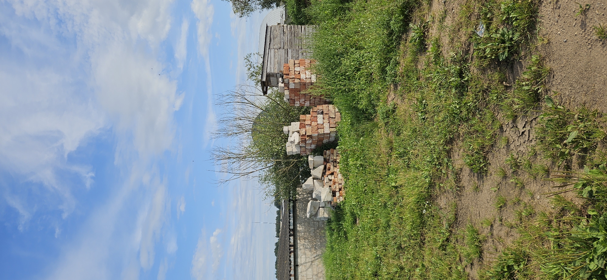 An exterior vertical shot captures a construction site in Мазалаўскі сельскі Савет, Belarus. The foreground is dominated by a patch of dry, cracked earth with sparse green grass. In the mid-ground, piles of bricks and concrete blocks are scattered around a cluster of vegetation. To the left, a low, long building with a grey, corrugated roof stands against a grey concrete wall. To the right, a small, weathered wooden structure with a slanted roof sits next to more stacks of bricks and lush green foliage. The background features more greenery and a large, light-colored domed structure partially obscured by trees. The sky is a vibrant blue with scattered white clouds.