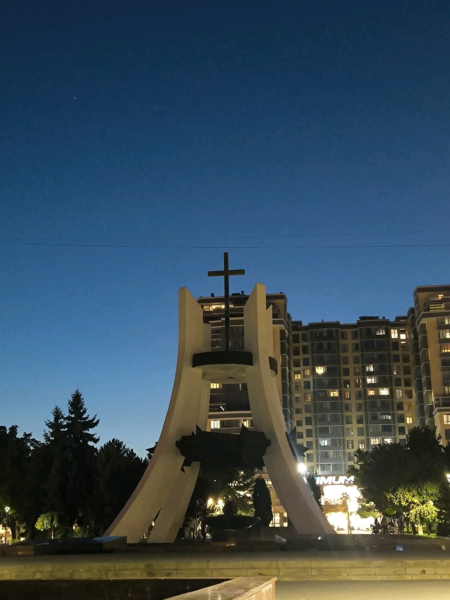 The image depicts the "Wings of Liberation" monument located in Chișinău, Moldova, at dusk. The monument, with its striking white, wing-like structures and a prominent cross at its apex, stands against a backdrop of a deep blue sky. To the right of the monument, a tall, multi-story apartment building with illuminated windows is visible, suggesting it is evening. Trees and park lighting are present around the base of the monument and in the surrounding area. A faint wire is stretched across the upper portion of the sky. In the sky, a single bright star is discernible. There are no people clearly visible in the frame.