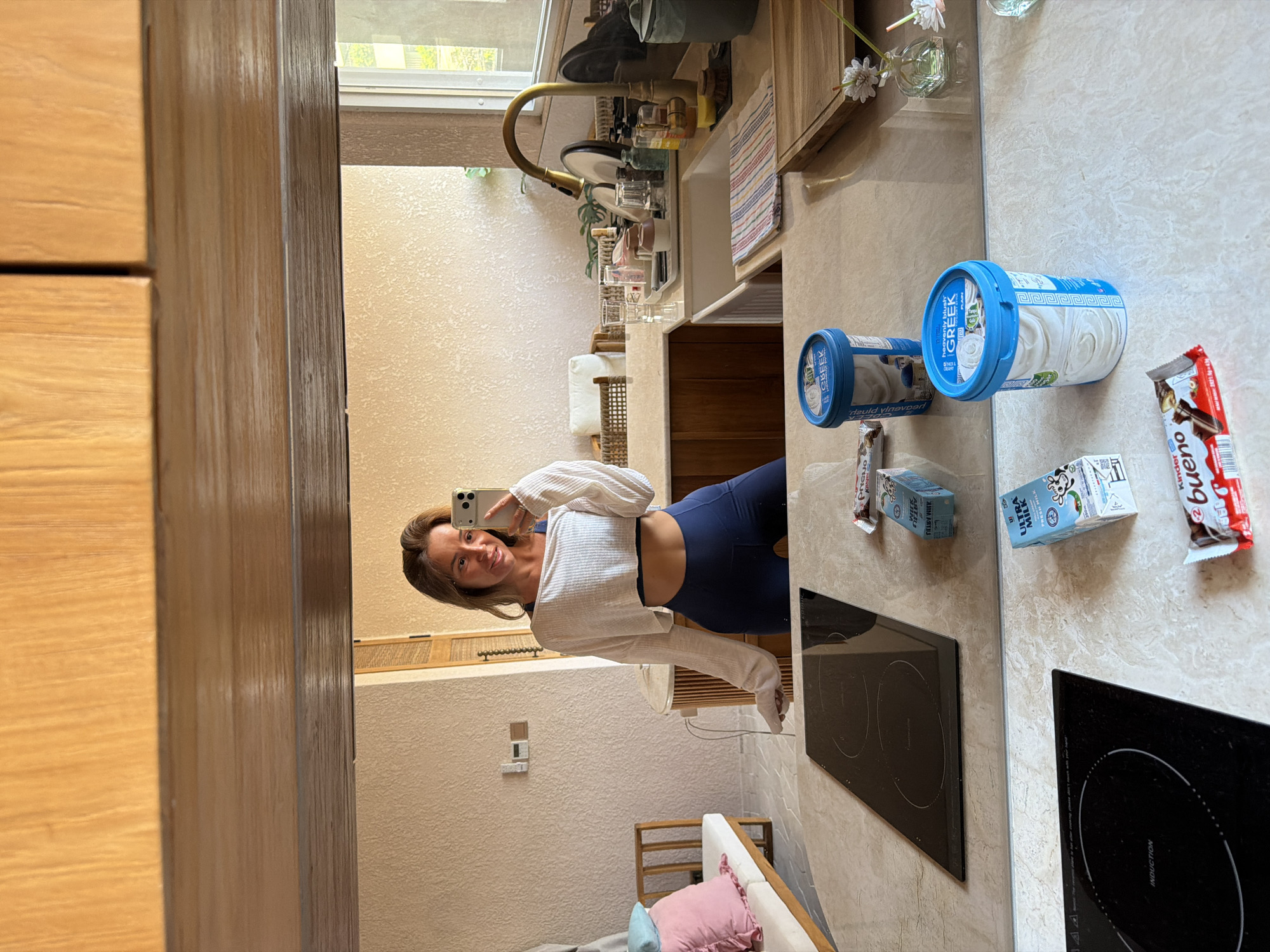 In a kitchen with light-colored walls and wooden cabinetry, a woman is taking a mirror selfie. She is wearing a white cropped long-sleeved shirt and navy blue leggings. She is holding a smartphone in her right hand. In the foreground of the image, on a marble countertop, are two containers of Greek yogurt, two small cartons of milk, and a Kinder Bueno chocolate bar. A black induction stovetop is visible on the left side of the countertop. In the background, a sink with dishes, a faucet, and a window overlooking a green outdoor area are visible. The scene appears to be set in a residential kitchen.