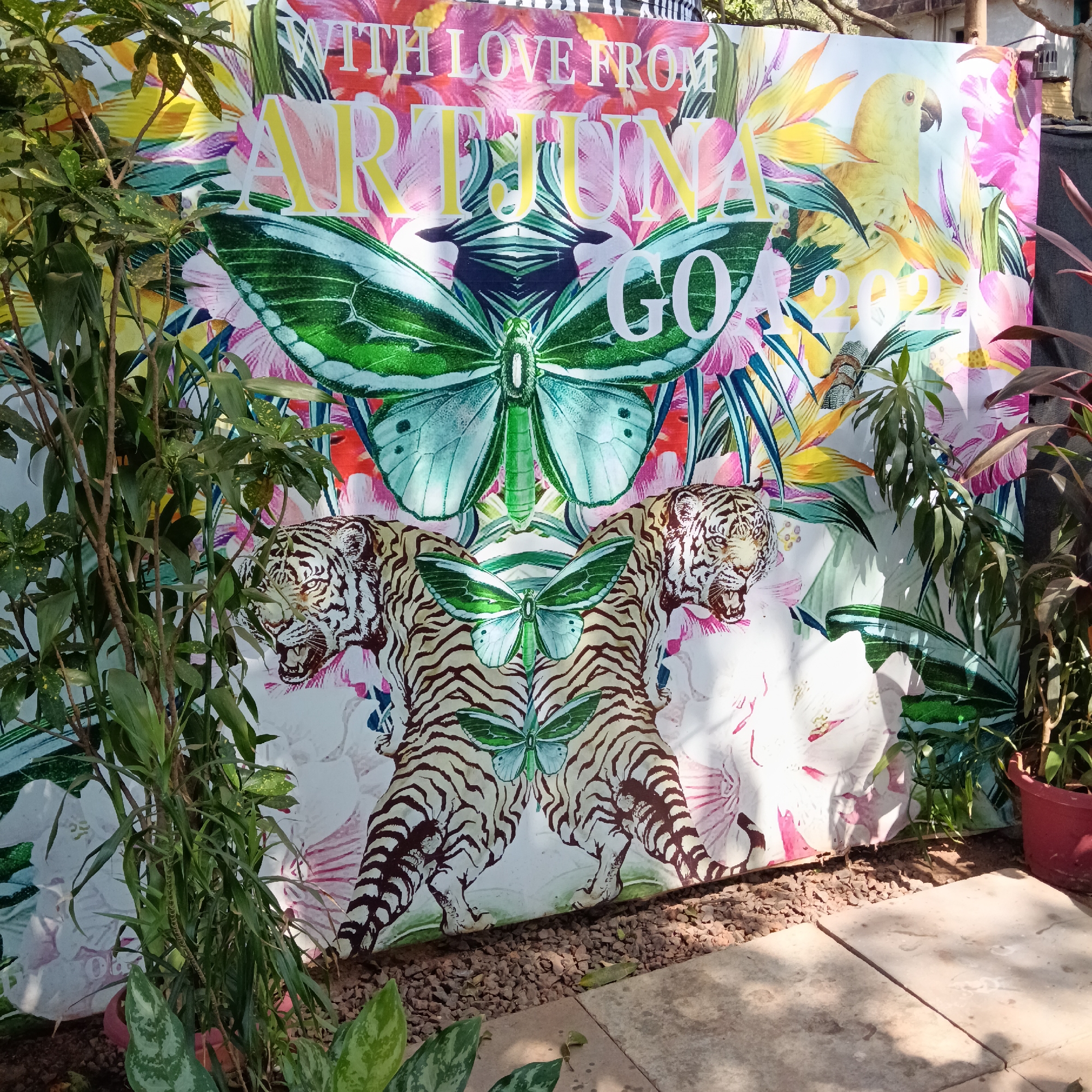 The image shows a colorful backdrop that reads "WITH LOVE FROM ARJUNNA GOA 2023," possibly a photo booth or decorative element. The backdrop features images of tropical flowers, a parrot, a large butterfly, and two tigers. The backdrop is partially obscured by leafy plants on either side. The setting appears to be outdoors, with small gravel covering the ground and concrete paving stones visible. This is likely located in Anjuna, Goa, India, as indicated by the text on the backdrop. There are no people in the frame.