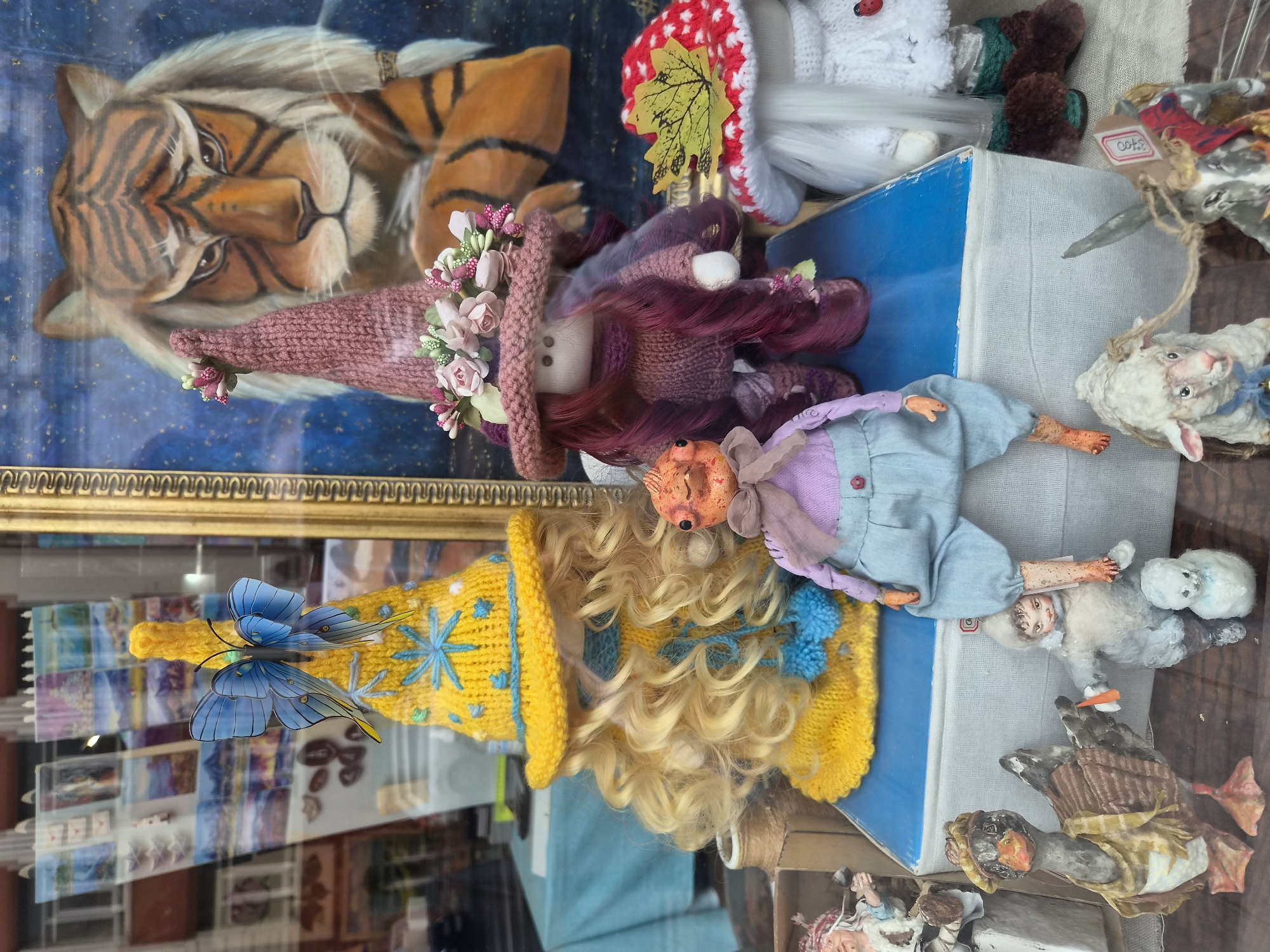 The image depicts a display of handmade dolls and figurines in what appears to be a shop window. In the foreground, to the left, a goose figurine wears a brown striped vest and yellow scarf. Next to it, a doll dressed in a white fluffy outfit holds a carrot and stands beside a small snowman figure. A sheep figurine with a blue ribbon is positioned to the right of the goose and doll. Further back, a grey donkey figurine with a price tag is visible.
Dominating the mid-ground are two larger doll figures. The one on the left wears a knitted yellow witch's hat adorned with a blue butterfly and has blonde curly hair. The doll on the right is dressed in a knitted purple outfit, a pink witch's hat decorated with flowers, and has long dark purple hair. Behind these dolls, a painting of a tiger's face is displayed. To the far right, a doll dressed as a mushroom, with a red and white spotted hat, is partially visible.
The background shows a shop interior with shelves and various items on display, including what look like postcards or small paintings. The entire scene is viewed through glass, with reflections of the street or building opposite visible on the surface. Based on the context of the provided additional hint, this display is located in Vladivostok, Russia.
