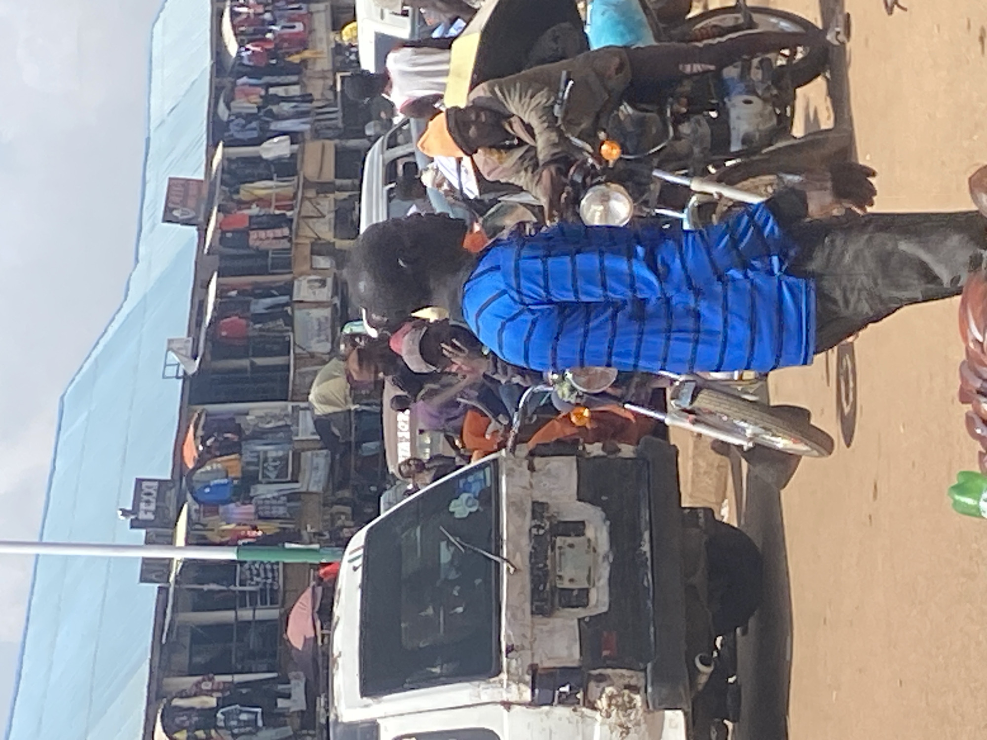 In the city of Bukuru, Nigeria, a bustling street scene unfolds with a focus on commerce and daily life. In the foreground, a Black man, wearing a blue and black striped long-sleeved shirt and dark pants, stands facing left, his body angled slightly towards the viewer. To his right, another Black man, wearing an orange beanie and a brown jacket over a lighter-colored garment, sits astride a motorcycle, looking towards the right. Behind them, a white van with significant damage to its rear is parked. The van appears to be old and weathered, with parts of its exterior missing or discolored.
The background reveals a vibrant marketplace with numerous stalls, displaying a variety of clothing items such as shirts, jackets, and trousers, hung out for sale. A light blue corrugated metal roof covers the market structures, suggesting an open-air or semi-enclosed market. Several other individuals are visible in the background, some standing, some seated, and some appearing to be engaged in transactions. Motorcycles and other vehicles are also present, indicating active transportation and trade. The ground is a dusty, reddish-brown dirt, typical of many Nigerian roads and market areas. The overall impression is one of a lively and active commercial hub.