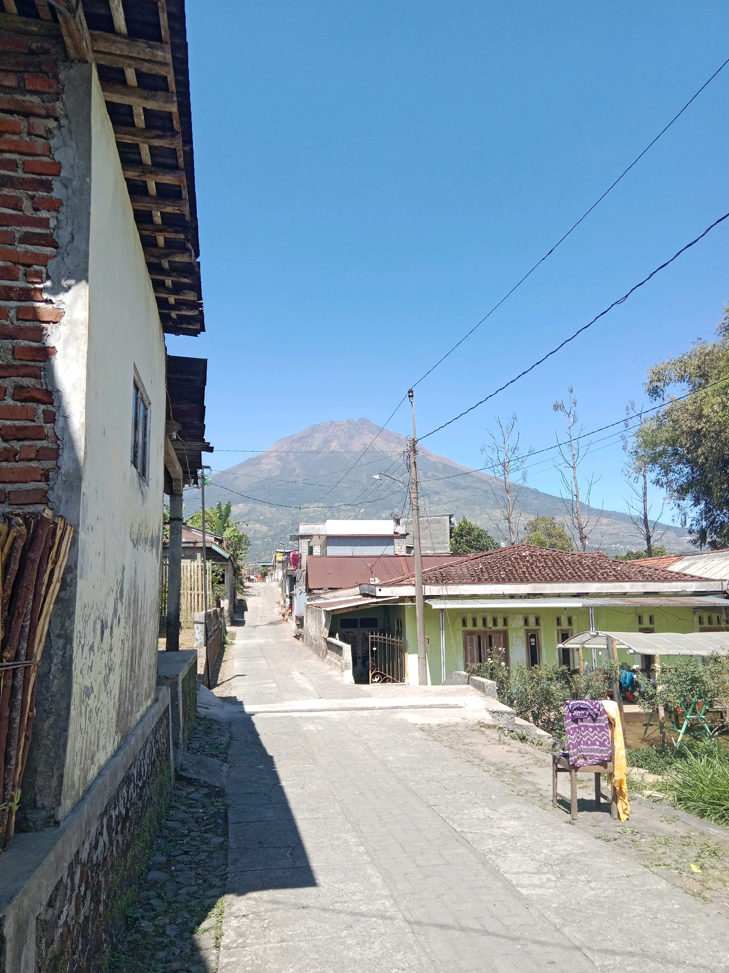 The image shows a view down a paved street in the city of Jurangjeru, Indonesia. The street is lined with houses on both sides. A large mountain can be seen in the background. The sky is clear and blue. On the left side of the street, there is a house with a brick wall and a white facade. On the right side of the street, there are several houses, one of which is painted light green. A wooden chair with clothes draped over it is placed on the right side of the street. There are power lines visible in the sky. The overall scene depicts a residential street in a tropical environment.