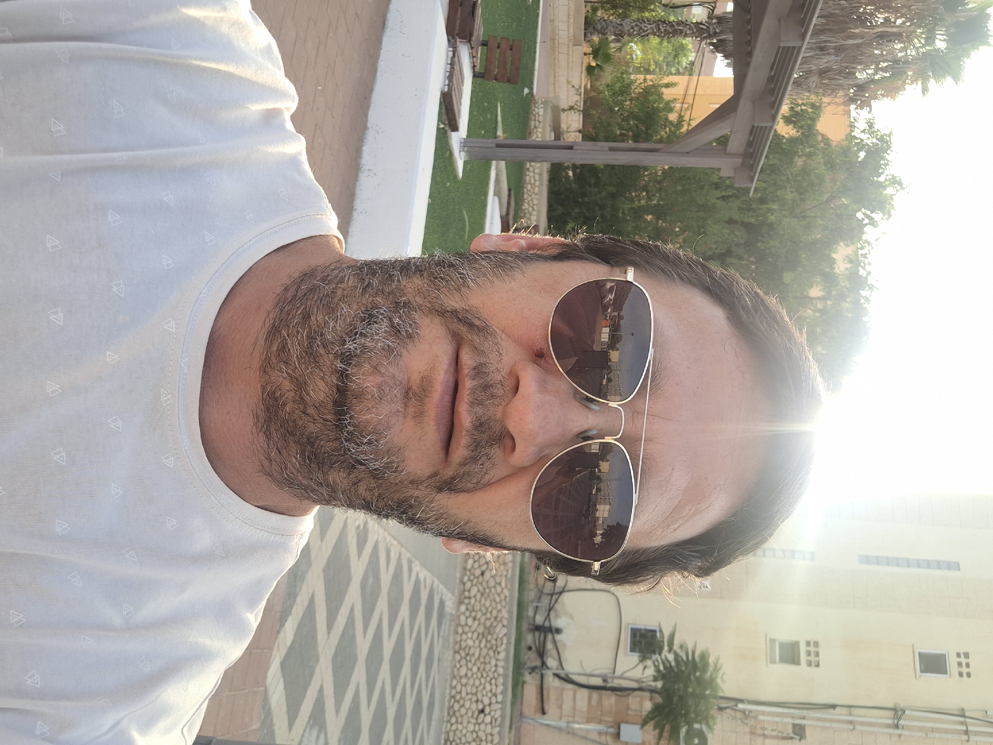 A selfie of a man with short dark hair, sunglasses, and a beard is taken outdoors in Yerocham, Israel. He is wearing a light-colored t-shirt with a triangular pattern. The man is looking directly at the camera. Behind him, there is a building with windows and a balcony, a wooden pergola with palm trees, and green grass with benches. The sun is shining brightly, creating lens flare. The ground in front of him is paved with interlocking bricks. The scene appears to be a public outdoor space, possibly a park or a courtyard.