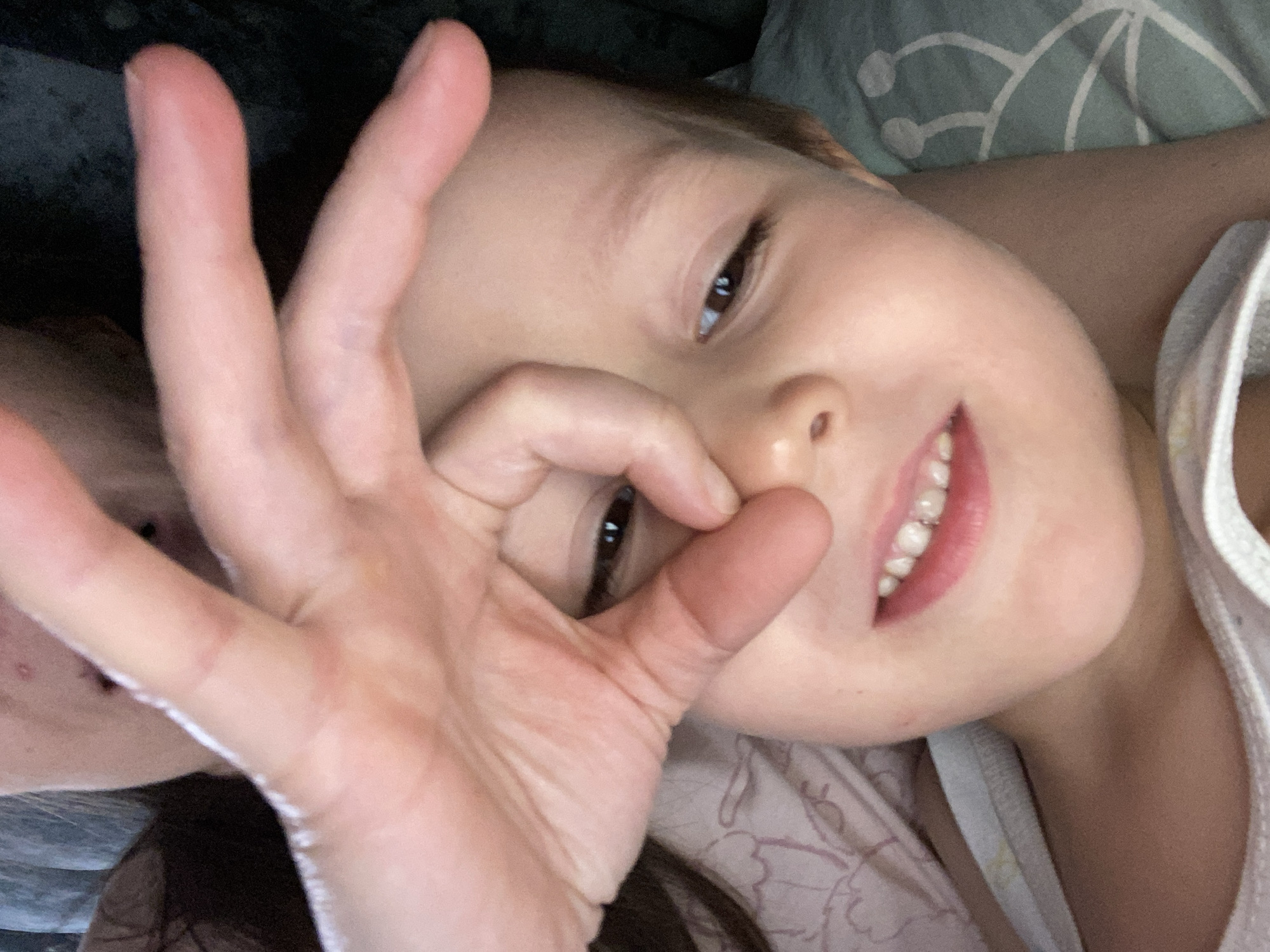 A child is lying down, looking at the camera, and making a circle with their thumb and index finger over their eye. They are smiling and wearing a white tank top. The background shows bedding. There is no indication of the location being Chaikovsky, Russia in the image.