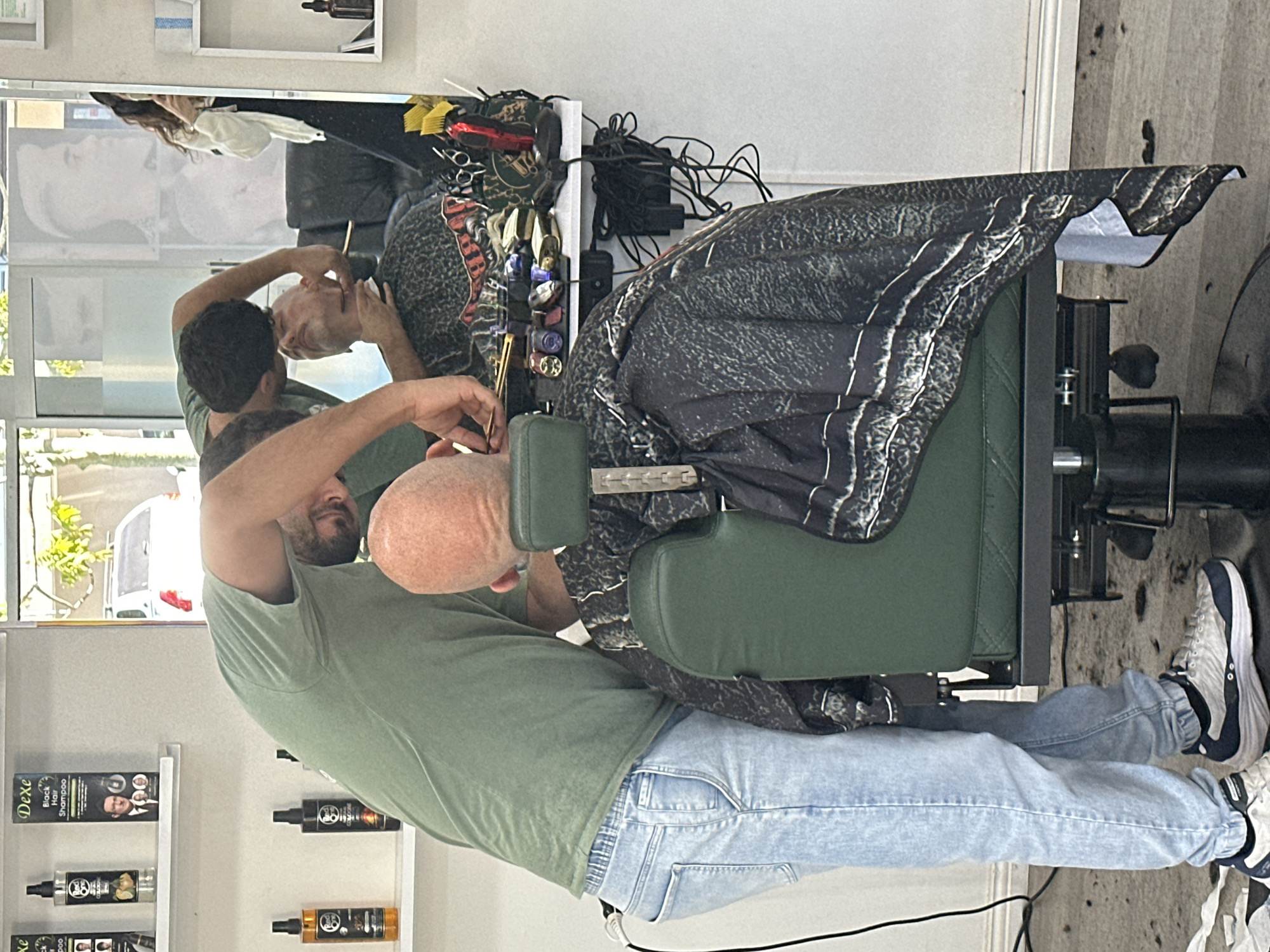 The image depicts a scene inside a barbershop, likely located in Coffs Harbour, Australia. A barber, wearing a green t-shirt and light-wash jeans, is leaning over a client who is seated in a dark green barber chair. The barber is holding a razor to the client's face, appearing to be in the process of shaving. The client, a bald man, is wearing a patterned cape. In the background, a mirror reflects another person, possibly a customer or staff member, holding a phone, and a faded image of a person with a haircut. Shelves are visible on the walls, displaying various hair products. The floor appears to be a light-colored wood or laminate. The overall environment suggests a professional grooming service.