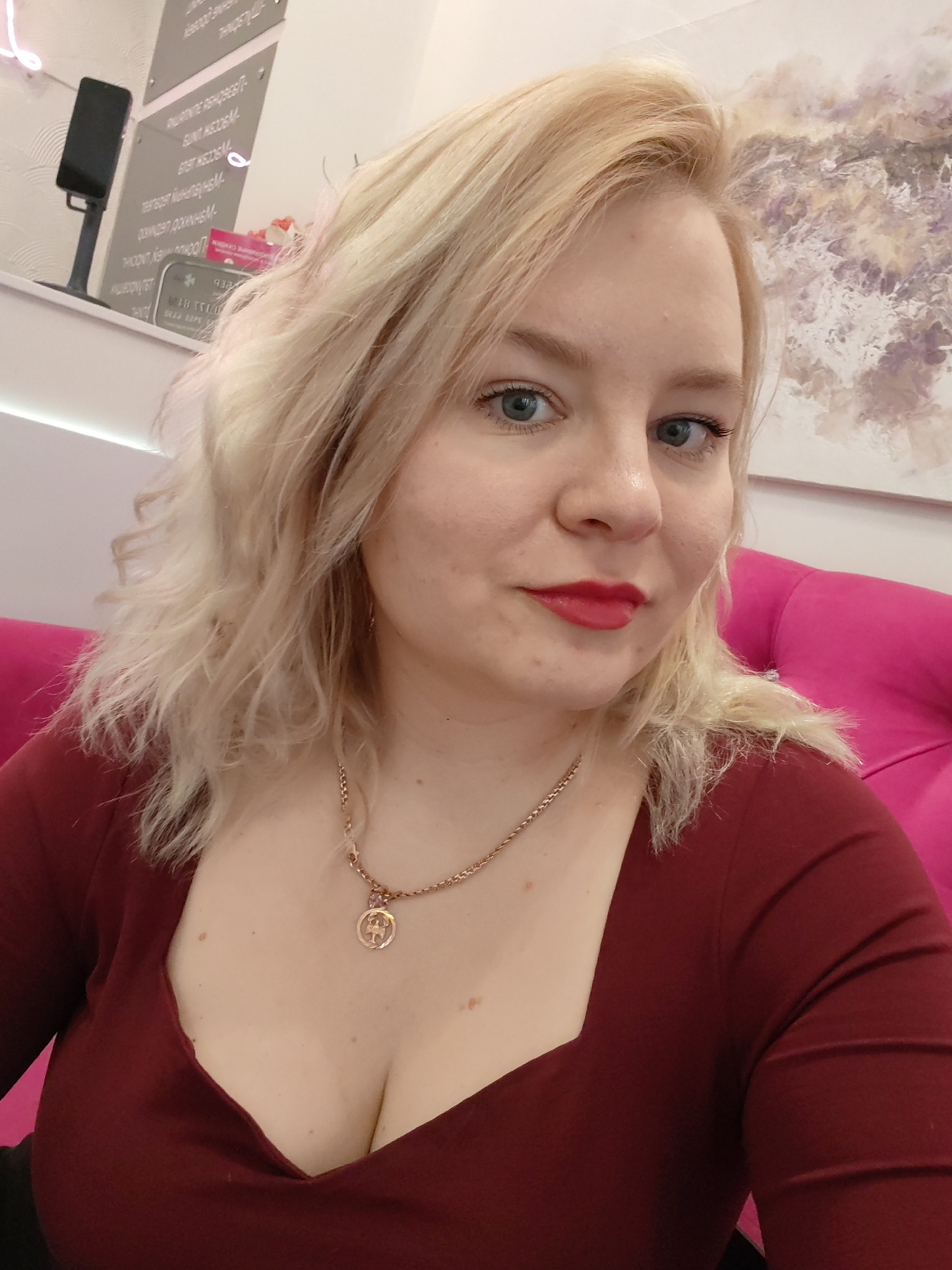 A portrait photograph captures a blonde woman from the chest up, looking directly at the camera with a calm, slightly smiling expression. She has fair skin, light blue-grey eyes, and is wearing red lipstick. Her medium-length blonde hair is styled with soft waves, falling over her shoulders. She is dressed in a dark red or maroon long-sleeved top with a square neckline, revealing some cleavage. A gold chain necklace with a round, detailed pendant, possibly depicting an angel or winged figure, rests on her chest.
The setting is indoors, featuring a modern and well-lit environment. To her right, a bright fuchsia-colored chair or sofa armrest is visible. The background includes a white wall adorned with various decorative elements. On the left, a black rectangular device on a stand is reflected in what appears to be a mirrored or reflective panel. This panel and others behind her display mirrored text in Cyrillic script, which, though blurry and difficult to read precisely due to reflections, appears to list services or information. Above the woman's head, an abstract painting with light purple and brownish hues adorns the wall. A thin, light strip (possibly neon) runs along a white ledge in the background. The overall impression is that of a salon, spa, or a modern public waiting area. The woman is posing for the photo, with a direct and composed gaze.