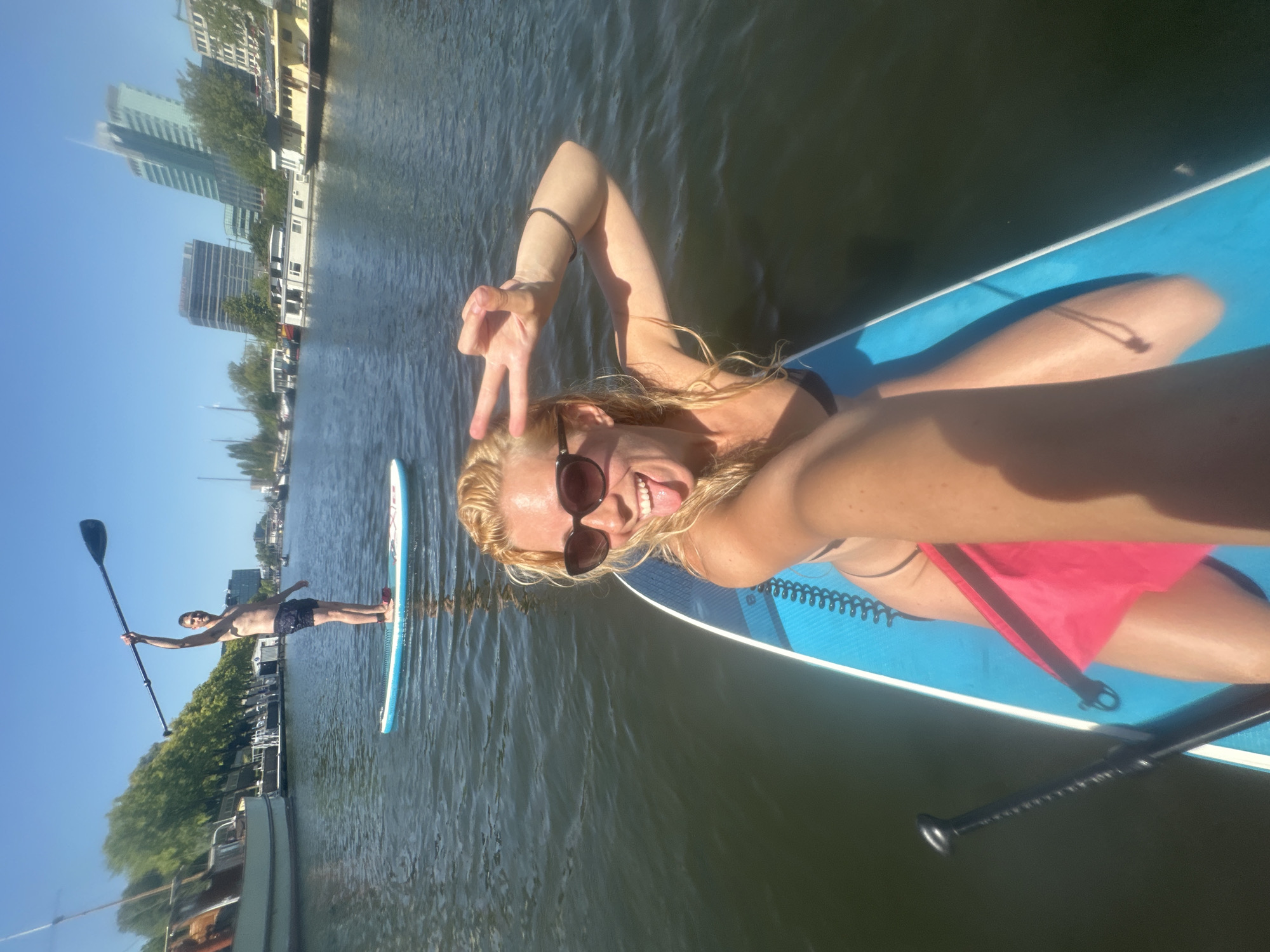 The image depicts two individuals stand-up paddleboarding on a canal in Amsterdam, Netherlands. In the foreground, a woman with blonde hair and sunglasses smiles and sticks her tongue out, holding up a peace sign with her right hand. She is on a blue paddleboard, wearing a black bikini top and a pink sarong is attached to the board. In the background, a man is stand-up paddleboarding further down the canal, holding his paddle aloft. The canal is lined with trees and buildings, including modern high-rises, and several houseboats are visible. The sky is clear and blue, indicating a sunny day.