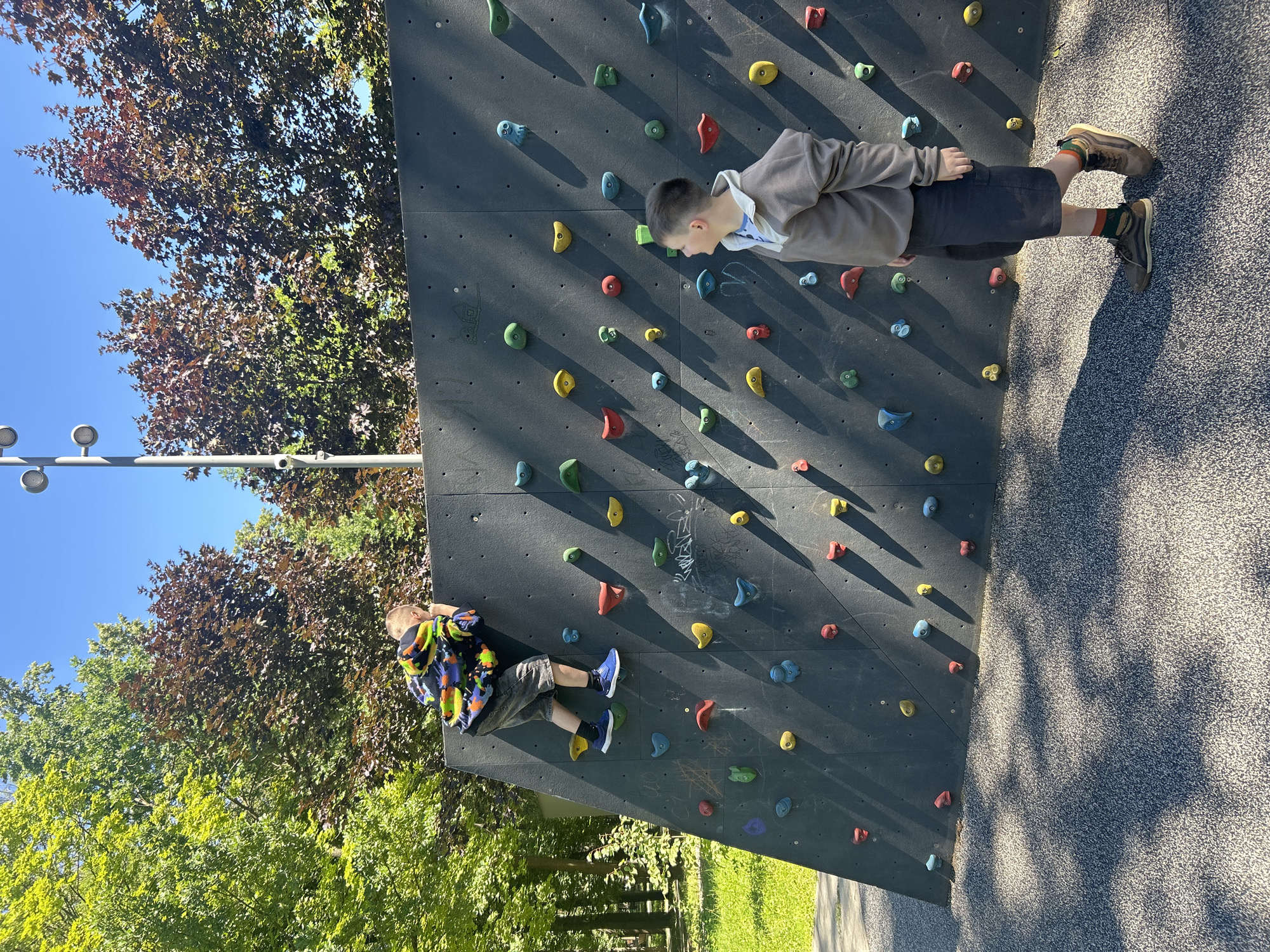 Two young boys are engaged in climbing a low, black climbing wall in an outdoor park. The boy on the left is actively climbing, positioned mid-wall, wearing a colorful patterned sweatshirt, grey shorts, and blue sneakers. The boy on the right stands at the base of the wall, looking up at the climber, dressed in a brown hooded sweatshirt, dark shorts, and brown shoes with distinctive socks. The climbing wall is studded with various colored handholds (red, yellow, blue, green). The park setting features lush green grass and mature trees with green and red foliage under a clear blue sky, suggesting a sunny day. The ground surface around the wall is a textured, speckled grey material. The scene is devoid of any explicit indicators of its specific location within Khimki, Russia, beyond its general urban park setting.