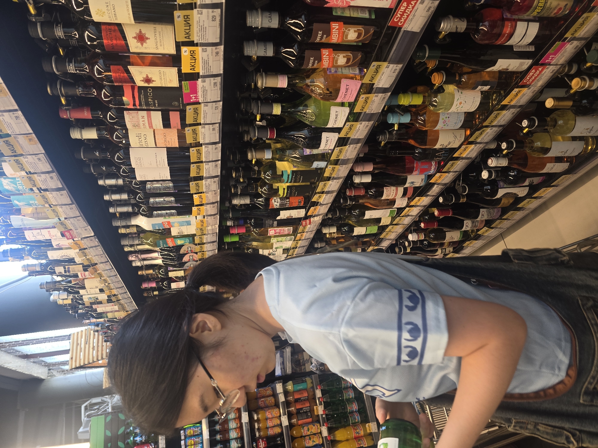 In the city of Ulan-Ude, Russia, a young woman with dark hair pulled back in a ponytail and wearing glasses stands in a supermarket aisle. She is wearing a light blue t-shirt with a blue pattern on the sleeve and a denim apron over dark pants. Her right hand holds a green bottle, and her left hand is near it. The background is filled with shelves stocked with various bottles of wine and other alcoholic beverages, many of which have yellow and red sale tags. The lighting suggests an indoor retail environment.