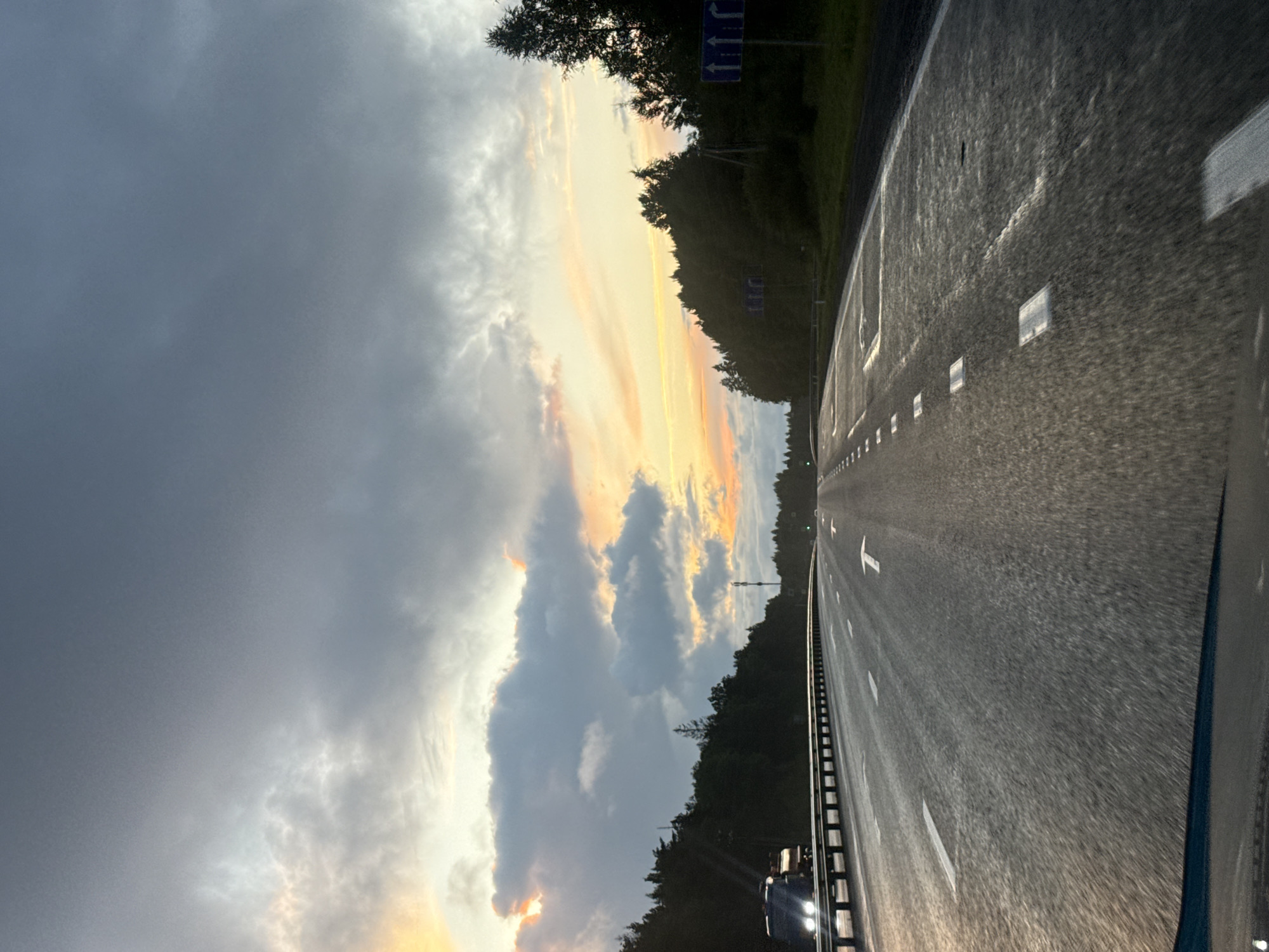 A wet highway cuts through a forested landscape under a cloudy sky during what appears to be sunset or sunrise. The road is marked with white lines, including directional arrows. A truck with illuminated headlights is visible in the distance on the left side of the road. Trees line both sides of the highway, with some appearing dense and dark. The sky is a mix of gray clouds and lighter, colorful hues of orange and yellow, suggesting a transition between day and night. Road signs are visible on the right side of the road, indicating traffic direction. The image is taken from the perspective of a vehicle traveling on the highway.