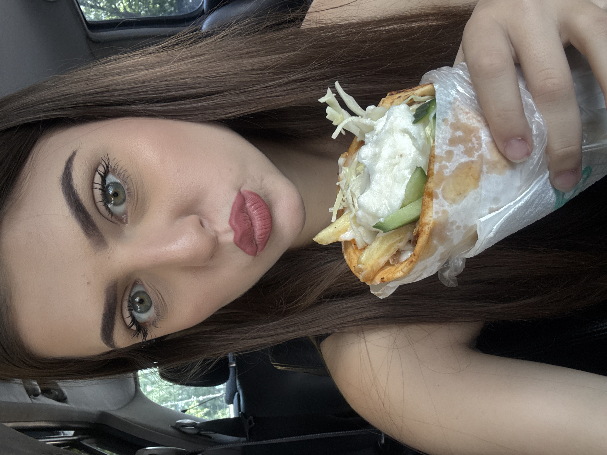 A woman is in the driver’s seat of a car and holding a gyro wrapped in paper. The gyro contains lettuce, cucumber, french fries, and a white sauce. The woman has brown hair, green eyes, and pink lipstick. The location appears to be Burgas, Bulgaria based on visual markers in the image.