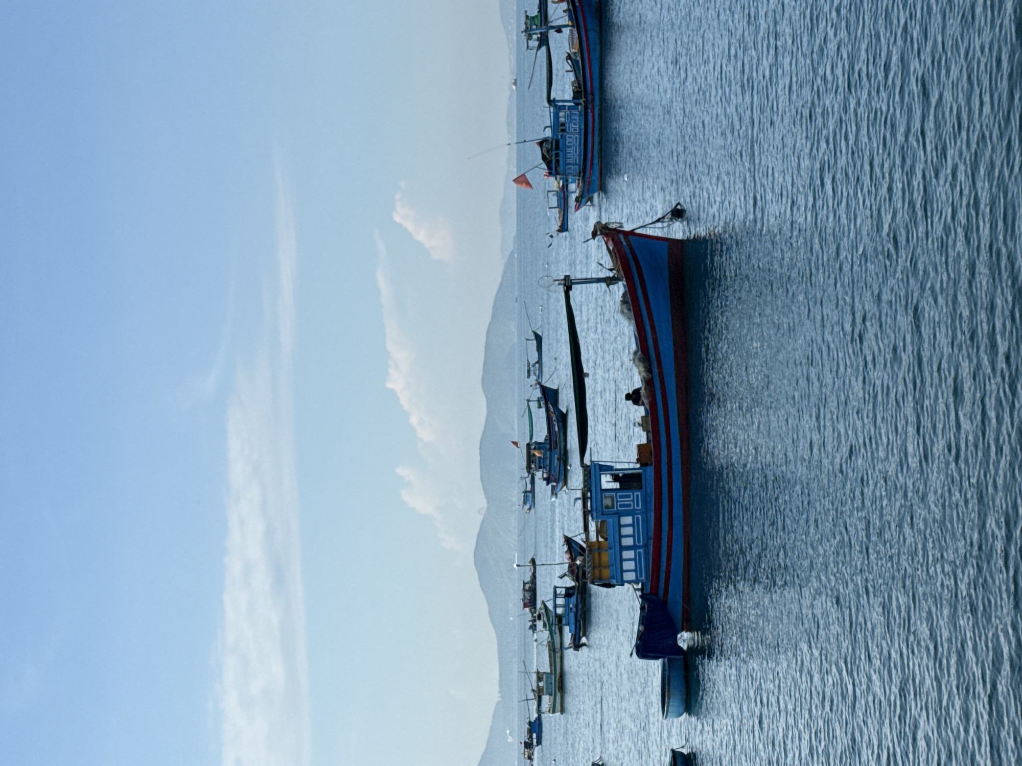 The image captures a harbor scene in Bac Nha Trang Ward, Vietnam, on a clear day with a blue sky and scattered white clouds. The water is calm and deep blue. Numerous fishing boats of various sizes are anchored, filling the frame. The most prominent boat in the foreground is a blue and red vessel with a cabin structure. A person is visible sitting in the cabin of this boat. Other boats are scattered throughout the scene, some are larger with multiple levels, while others are smaller and simpler. In the background, a hazy mountain range forms the horizon. The overall impression is one of a working fishing harbor, with boats resting in the water under a bright sky.