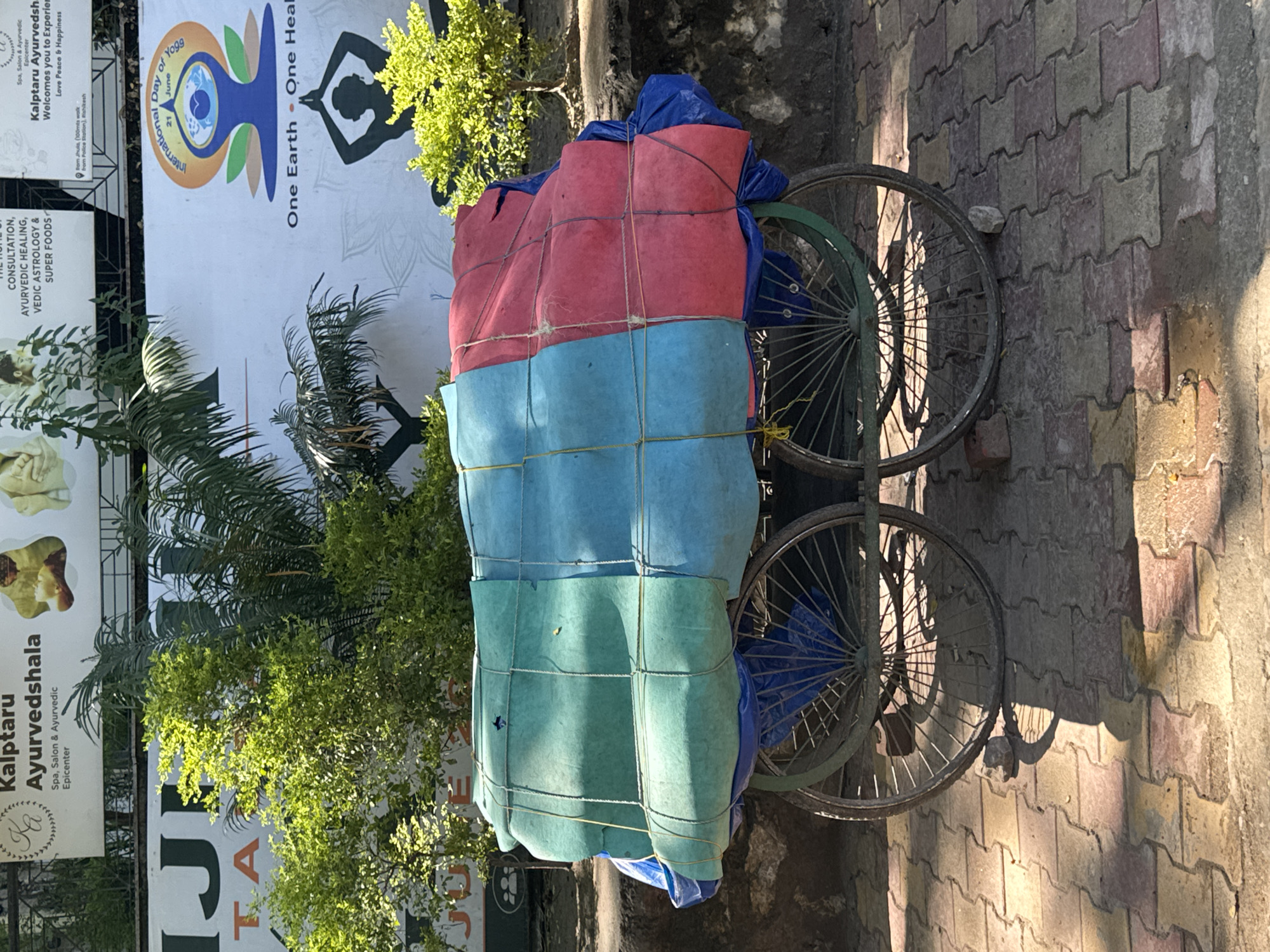 The image captures a street scene in Rishikesh, India, focusing on a two-wheeled cart heavily laden with goods covered by colorful tarps. The cart, a common sight for transporting merchandise, is positioned on a paved walkway composed of interlocking bricks. The tarps are a combination of green, blue, and red, secured with rope. The cart's wheels are visible, one more prominently than the other, showing a radial spoke design. Behind the cart, a blurred background includes greenery from trees and foliage, suggesting an outdoor setting. To the right, a portion of a billboard is visible, advertising "International Day of Yoga" with imagery related to yoga and wellness, also mentioning "One Earth • One Health." The overall lighting indicates a sunny day, casting shadows from the cart and vegetation onto the pavement. The scene appears to be an ordinary day in Rishikesh, likely a commercial or residential area where such carts are utilized for transportation and commerce.