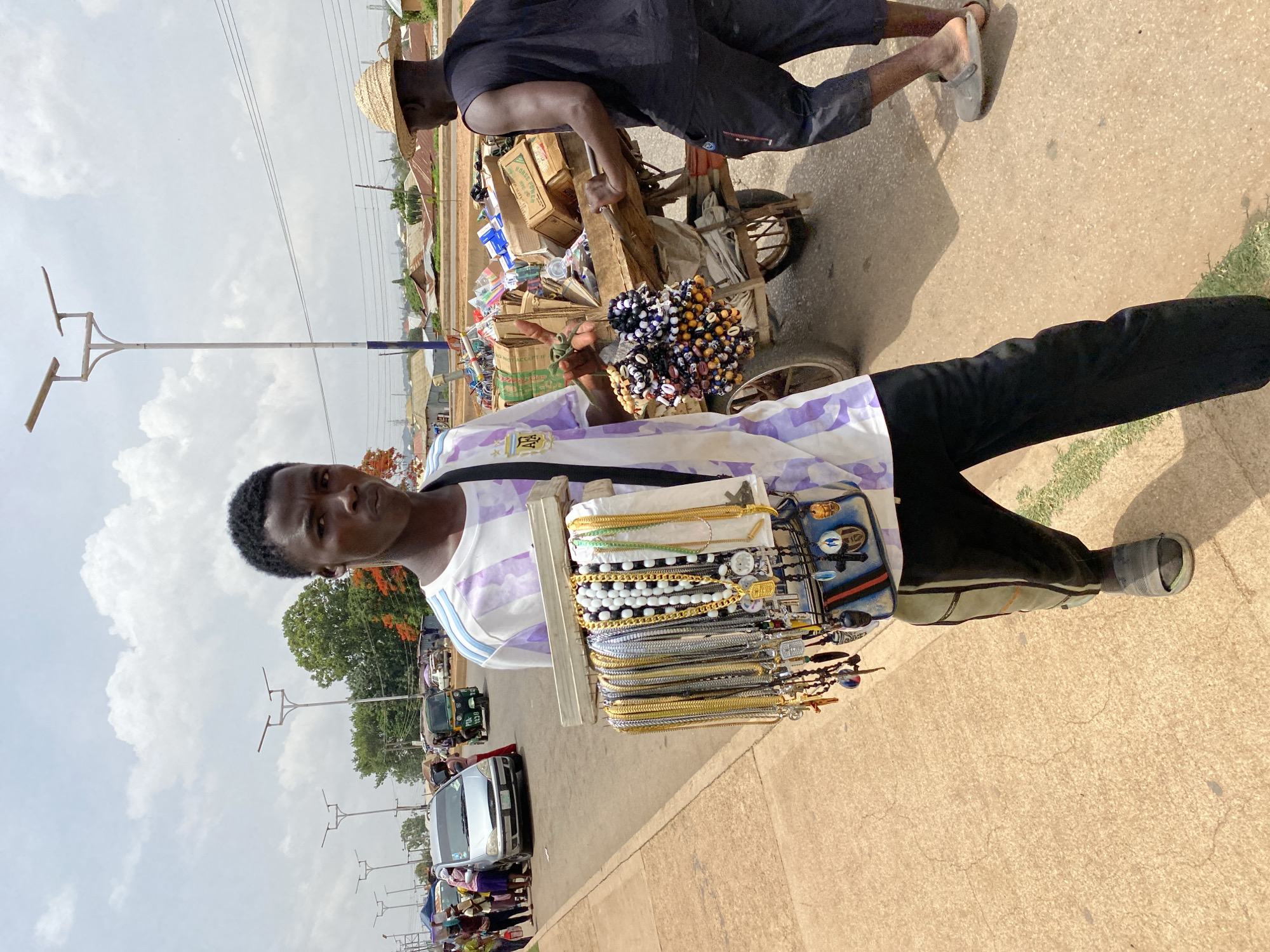 The image depicts a street scene in Bukuru, Nigeria. A man wearing a white and light purple striped shirt with the Argentinian flag logo on the left breast, along with black trousers and grey shoes, stands facing the camera, displaying an assortment of jewelry hung around his neck. The jewelry includes gold and silver-colored necklaces, bracelets, and wristwatches.
In the background and further along the same road, another man wearing a black top and trousers with a straw hat is pushing a wooden cart loaded with assorted goods and more jewelry. Cars and three-wheeled vehicles, known locally as “Keke Napep,” are also visible on the road, suggesting traffic. Street lights line the roadside. The ground appears to be a paved road or sidewalk, with some grass visible along the edge. The sky is partly cloudy.