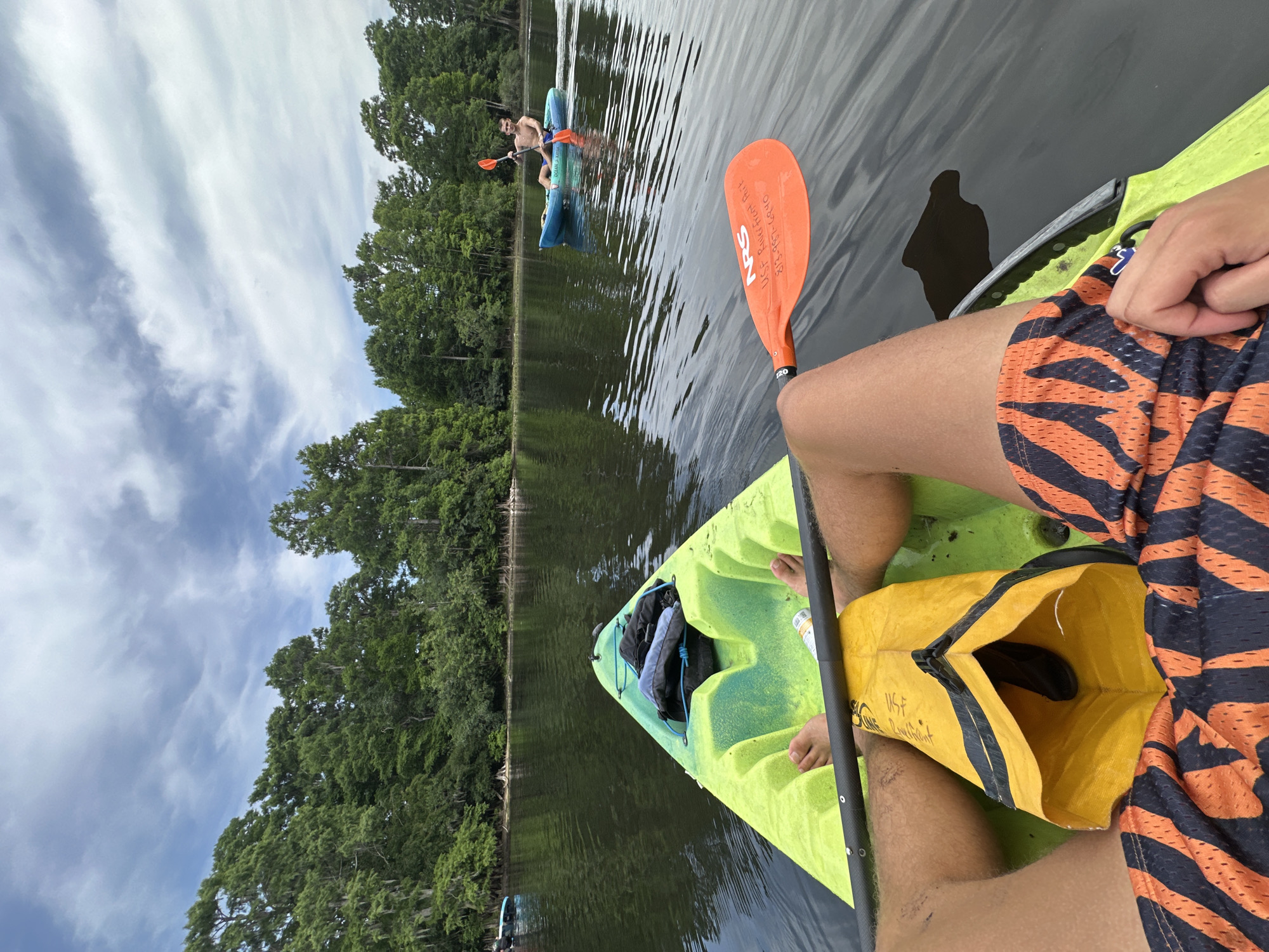 This image, presented from a first-person perspective, captures a serene daytime kayaking scene on a calm body of water, likely a river or lake. The viewpoint is from inside a bright lime green kayak, looking forward and slightly towards the left.
In the foreground, the kayaker's legs are visible, clad in orange and black patterned swim shorts. An orange paddle, bearing the text "NRS", and handwritten details "USF Riverfront Park" and "337-639-5840", rests across the kayak. Between the kayaker's legs, a yellow dry bag is open, showing "USF Riverfront" partially written on it, and containing a dark object. The front of the green kayak holds what appears to be a grey bag or backpack.
Further ahead on the water, another person is seen paddling a light blue kayak. This individual is shirtless and faces partially towards the camera. The water is dark and still, providing reflections of the surrounding environment.
The banks of the water body are densely lined with tall, vibrant green trees, creating a natural and lush backdrop. The sky above is partly cloudy with streaks of white and grey clouds against a light blue, indicating fair weather. The overall ambiance suggests a peaceful and leisurely outdoor activity. The image is oriented vertically, rotated 90 degrees counter-clockwise from its horizontal capture.