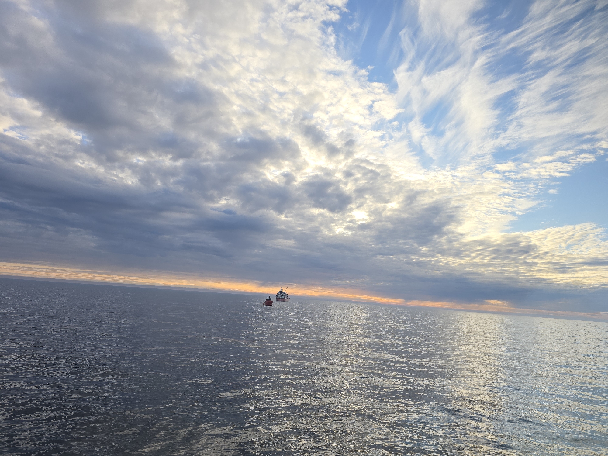 The image depicts a serene seascape under a sky filled with scattered clouds. The water stretches to the horizon, its surface reflecting the light. Two vessels are visible: one appears to be a larger ship, while the other is a smaller, likely a tugboat. The horizon line is faintly tinged with an orange hue, suggesting either sunrise or sunset. No specific location within the city of Unknown, Unknown can be determined from the image.