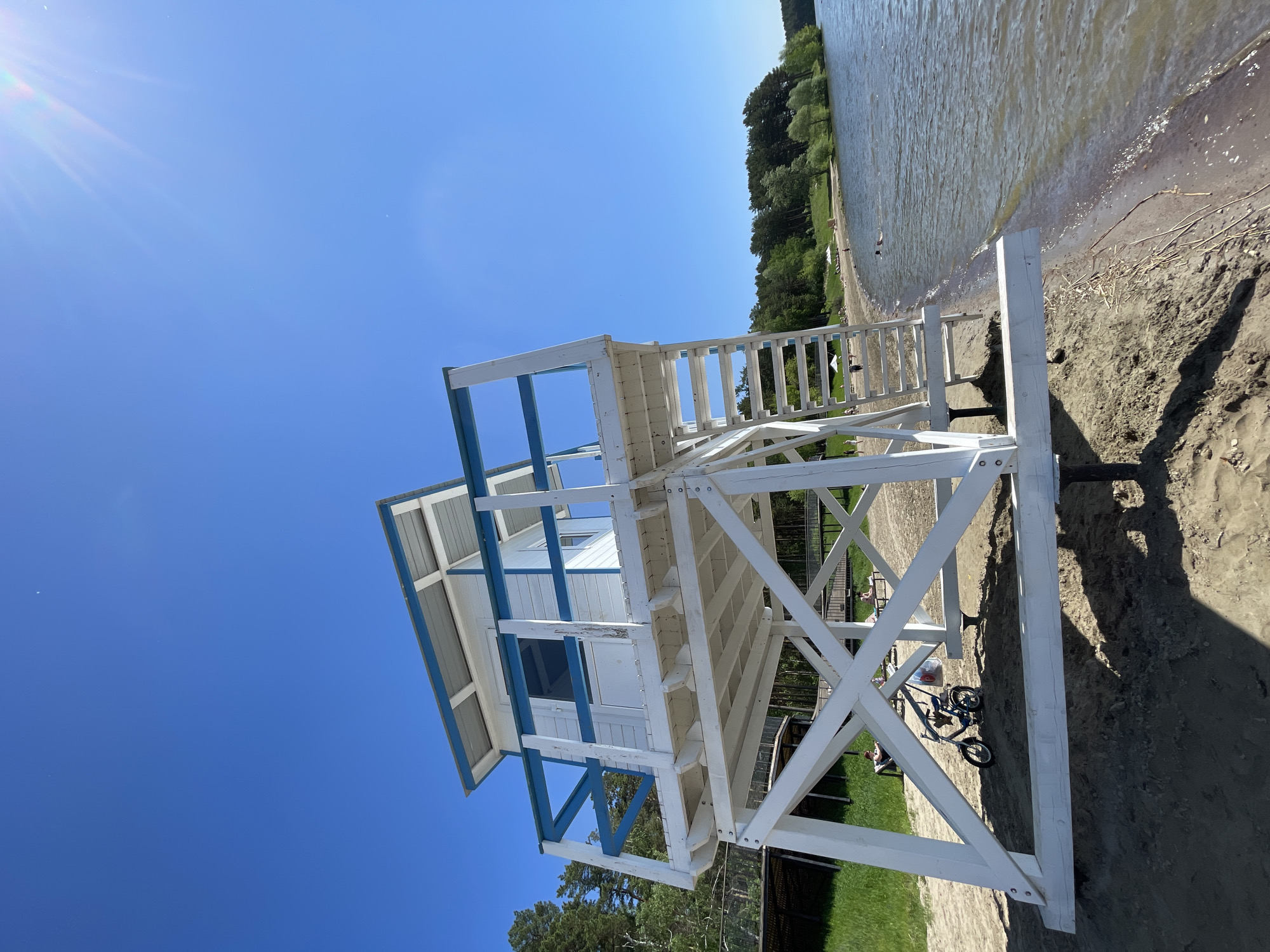The image depicts a bright, sunny day at a beach or lakeside area in Zheleznogorsk, Russia. A prominent white wooden lifeguard stand with blue trim occupies the left and central portion of the frame. The stand features an enclosed cabin at its upper level and stairs leading up, supported by a robust framework of white wooden beams, including clear cross-bracing.
To the right of the lifeguard stand, a wide body of water, likely a lake, extends across the scene. Small waves gently lap against the sandy shore, and the water appears a muted greenish-brown near the edge. The foreground consists of sandy beach, which gives way to a strip of green grass inland, visible beneath and behind the stand. In the distance, a dense line of green trees borders the far bank of the water.
The sky above is a clear, brilliant blue, devoid of clouds, indicating excellent weather. A bright sun flare is visible in the upper left corner, confirming the high position of the sun. The strong sunlight creates distinct, dark shadows from the lifeguard stand cast onto the sandy ground.
Several small, distant figures are visible: one person is seated on the grass near the base of the lifeguard stand, and others are scattered further along the beach and grassy areas. A child's bicycle or scooter is parked on the sand near the stand's foundation, next to a small container. The scene evokes a peaceful and pleasant summer day at a recreational waterfront. There is no visible text in the image.