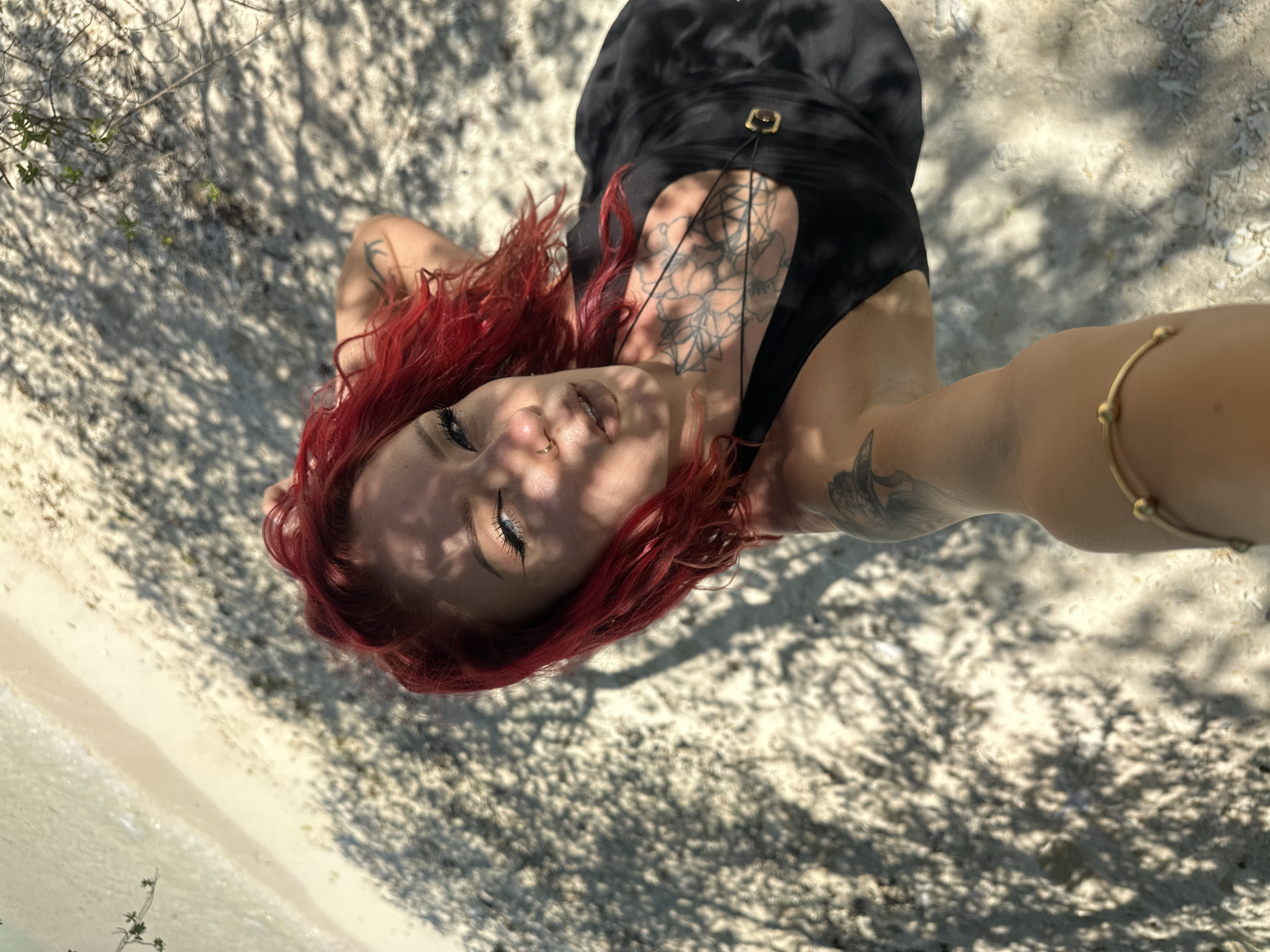 A young woman with bright red hair and several visible tattoos lies on a sandy beach, possibly near a body of water, under the partial shade of trees. She wears a black garment, possibly a dress or swimsuit, and a gold-colored bracelet is visible on her extended arm. Her face is turned towards the camera, and she appears to be looking upwards. The sand is light in color, and the shade cast by the trees creates a dappled light effect. The location is Unknown, Indonesia.