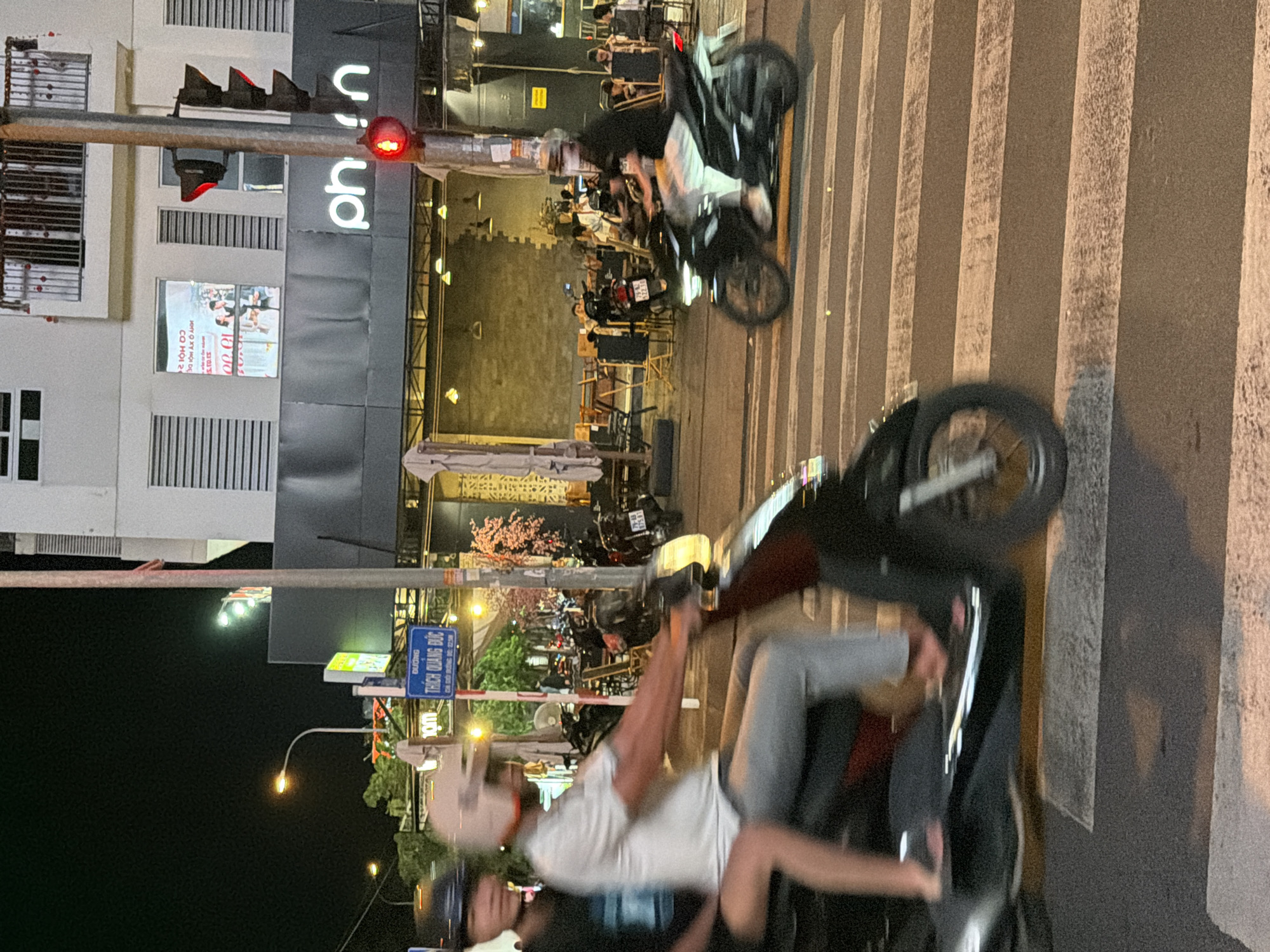 A night scene shows a busy street in Nha Trang, Vietnam, with several motorbikes on a zebra crossing. A man and a woman are riding on one motorbike, with the man driving and the woman as a passenger. The man is wearing a white helmet and a white t-shirt, and the woman is wearing a black helmet and a black t-shirt. In the background, there are several shops and restaurants with outdoor seating. The traffic light is red, indicating that vehicles should stop. The overall atmosphere is lively and energetic, with the presence of streetlights and neon signs adding to the vibrant nightlife of the city.