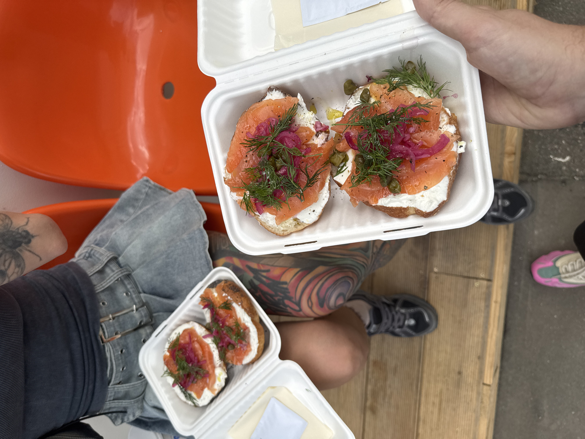 The image shows a top-down view of two open styrofoam containers holding open-faced sandwiches, likely lox bagels. Each bagel half is topped with cream cheese, salmon, red onions, dill, and capers. One container sits on a person's lap, whose leg is visible with a tattoo of swirling colors. They are wearing light-wash denim jeans with a black shirt. The person is sitting on an orange stadium-style seat. The other container is being held up by another person. To the right, a foot wearing a black and grey sneaker, and another foot wearing a white, pink, and green sneaker are visible on what appears to be a wooden platform. The image doesn't contain enough information to determine if the location is in Moscow, Russia.