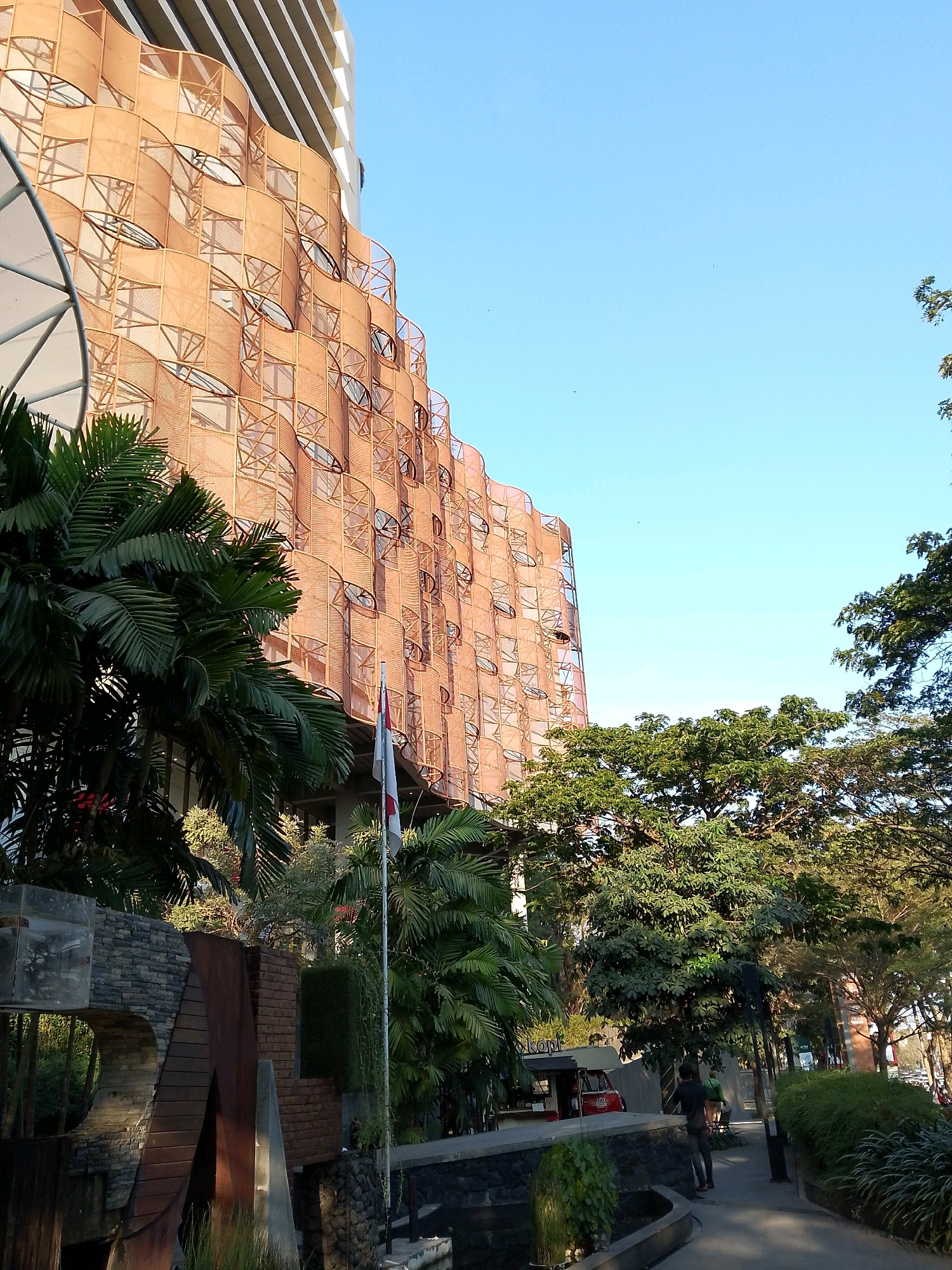 The image depicts the exterior of a building with an unusual, wavy, patterned facade in the city of Makassar, Indonesia. The building has a light-colored, multi-story structure behind the ornate outer layer, which appears to be made of interconnected panels or screens in a warm, orange-brown hue. These panels are arranged in a repetitive, curved pattern that follows the building's contour. Each panel contains a roughly oval-shaped opening, some of which appear to have glass or a reflective surface. A large palm tree with dark green fronds is prominently featured in the foreground on the left side of the image, partially obscuring the lower part of the building.
In the middle ground, a flag with red and white bands is visible on a flagpole, positioned in front of the building. Further back, dense foliage and trees create a lush, green environment, suggesting a tropical or subtropical setting. A paved pathway or sidewalk runs along the right side of the image, leading into the distance. A person, silhouetted against the sunlight, is walking away from the camera on this pathway. A small red food stall or truck is parked to the left of the pathway, near the building's entrance area. The sky is clear and blue, indicating fair weather. The overall scene suggests a modern architectural design integrated with natural landscaping in an urban environment.