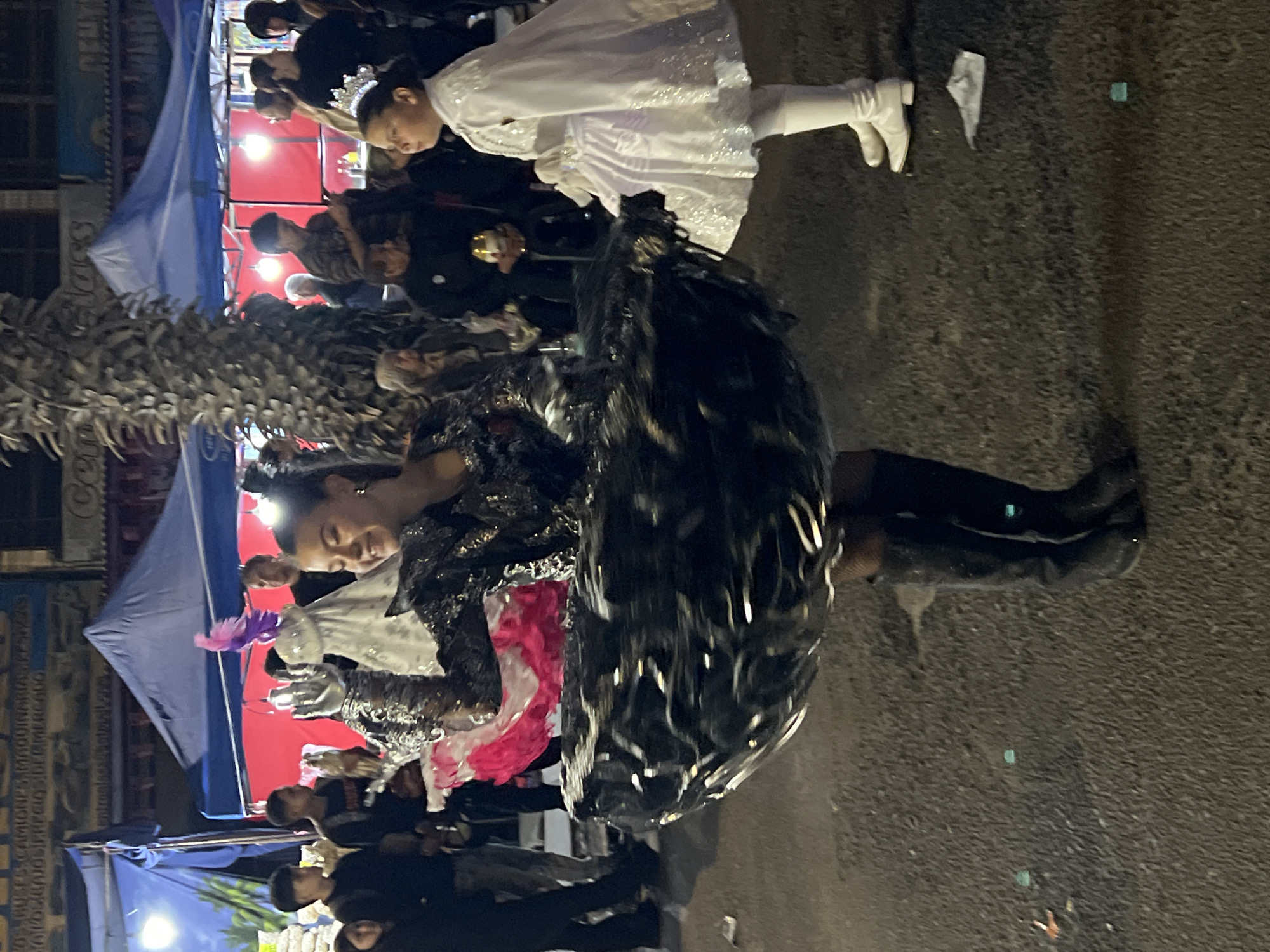 The image depicts a street scene at night in Arica, Chile, likely during a festive event or parade. In the foreground, a young woman wearing a dark, sparkling costume with a voluminous, ruffled skirt is the central figure. Her costume includes knee-high black boots and fishnet stockings. She is looking towards her left, with her right arm raised and hand outstretched.
To her right, a young girl in a white dress and cape, adorned with a tiara, stands looking to her left. She appears to be part of the same event.
In the background, several tents are set up, suggesting a market or stalls. People are milling around, some observing the costumed individuals. The street appears to be paved with asphalt, and there is debris scattered on the ground. The lighting suggests artificial illumination from the tents and possibly streetlights, creating shadows and highlighting the figures. The overall atmosphere is one of a community gathering or celebration.