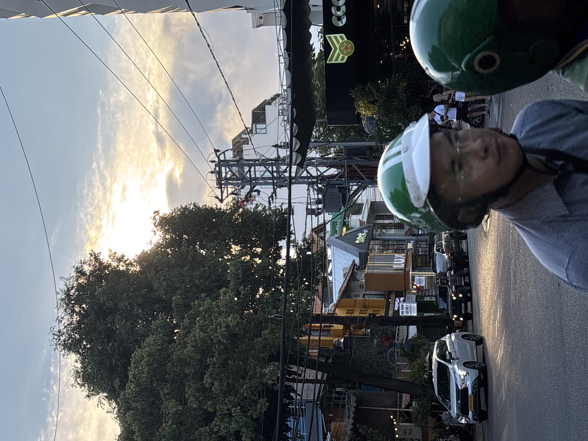 This media file captures a street scene in Nha Trang Ward, Vietnam, likely during the late afternoon or early evening due to the soft, diffused sunlight. The image is oriented vertically, with the sky and tree canopy dominating the left side, and the street with buildings and vehicles on the right.
The foreground features two individuals wearing green helmets, suggesting they are on a motorcycle. One person, likely the driver, is in focus, wearing a blue shirt and glasses, and their face is partially visible. The other person, further back, is mostly obscured by their helmet.
The background reveals a busy street lined with multi-story buildings. Some buildings have signs with visible text, including what appears to be Vietnamese characters. One prominent sign on a dark background has a green and red logo with a star. The street is paved and shows signs of traffic, with several vehicles including a white SUV and other cars and motorcycles parked or driving. A large, leafy tree provides shade on the left side of the street, with electrical wires crisscrossing the scene, typical of urban infrastructure in some regions. The sky is partly cloudy, with the sun partially visible through the clouds, creating a bright, atmospheric light. The overall impression is a snapshot of everyday life in a Vietnamese urban environment.