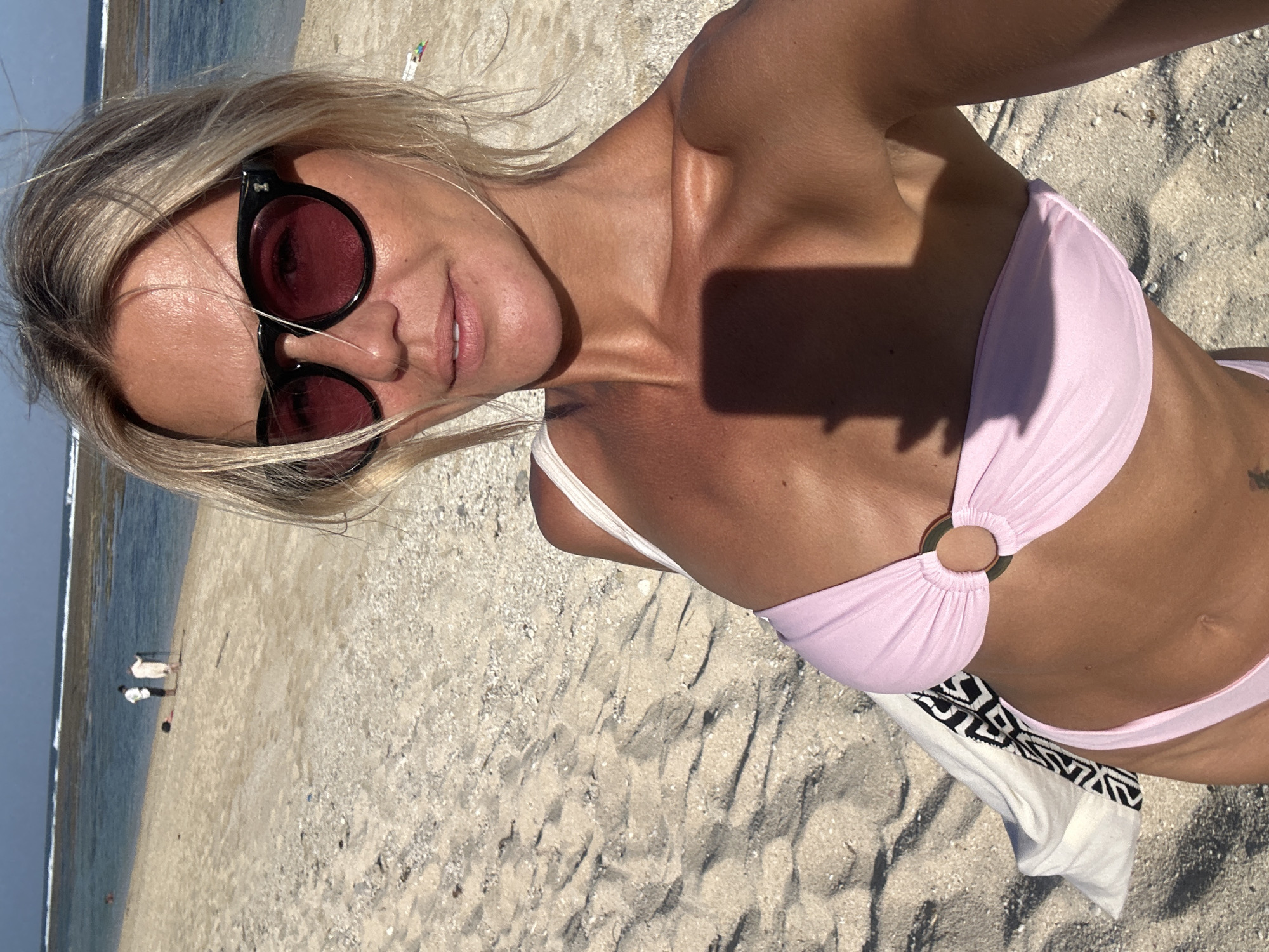 A woman with blonde hair and sunglasses takes a selfie on a beach in Kuta Selatan, Indonesia. She is wearing a pink bikini and a white bag with a black and white pattern is draped over her shoulder. The beach has light-colored sand and the ocean is visible in the background with gentle waves. Two people are standing near the water's edge in the distance. The lighting suggests it is daytime.