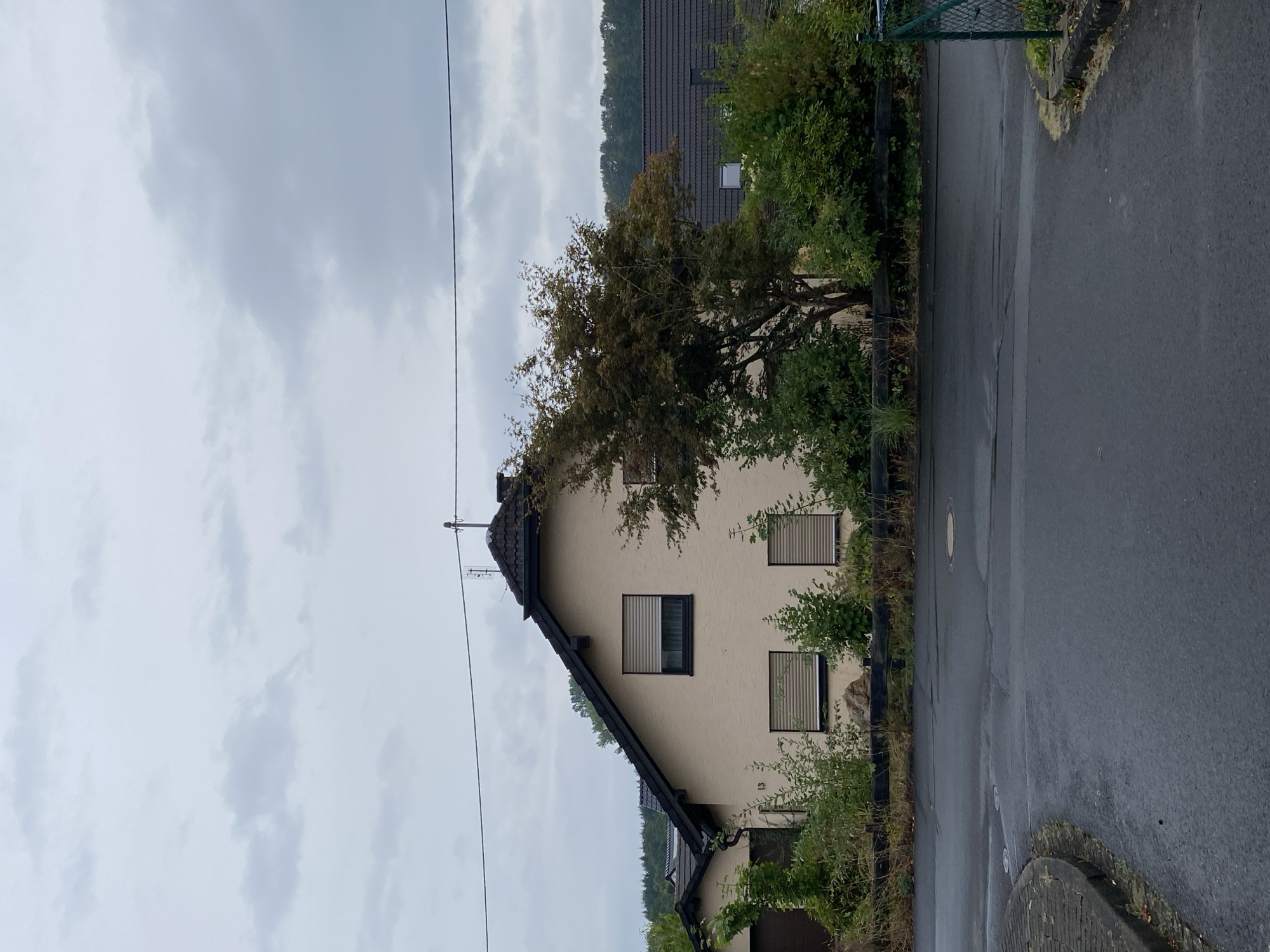 The image shows a detached house in Flammersfeld, Germany, on a cloudy day. The house is light beige with a dark tiled roof and black trim around the windows. There are roller blinds on the windows. A mature tree with dark foliage stands in front of the house, partially obscuring it. The property is bordered by a dark wooden fence or retaining wall, behind which is dense green shrubbery and some taller grass. The street in the foreground is asphalt and appears wet, suggesting recent rain. To the left, a brick-paved sidewalk curves away. In the background, more rooftops and dense trees are visible. A green chain-link fence runs along the right edge of the image. The house number "13" is visible on the facade.