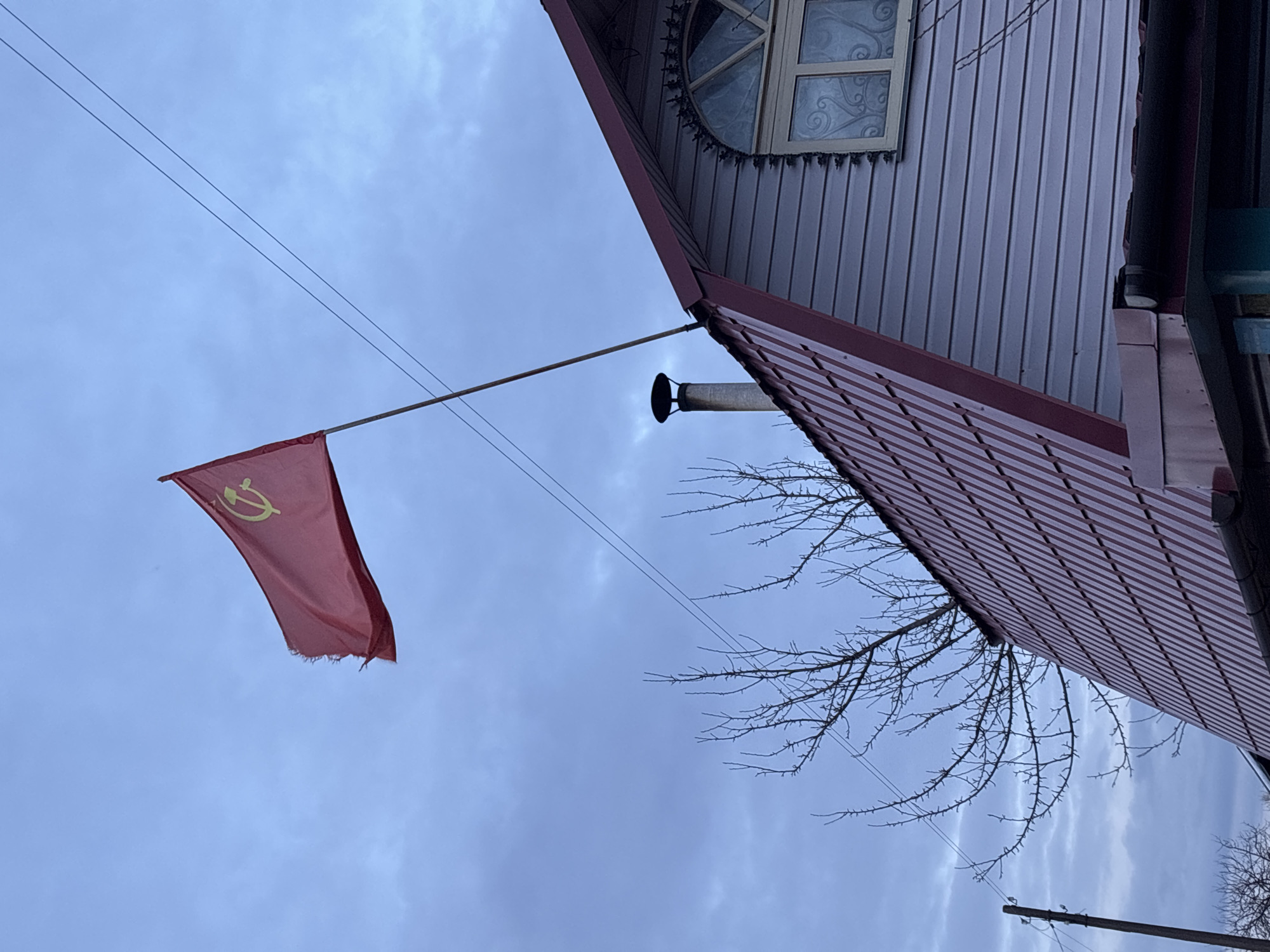 The image shows a tattered red flag bearing the yellow hammer and sickle, emblematic of the former Soviet Union, flying from a pole attached to the side of a house. The house has light-colored siding and a red roof. A window is visible on the upper portion of the house with decorative latticework. Power lines run across the upper portion of the frame. Bare tree branches are also visible. The sky is overcast. The flag's symbolism suggests a location within a former Soviet state. Based on the request for location specifics, it can be said the scene is likely in Chaikovsky, Russia.