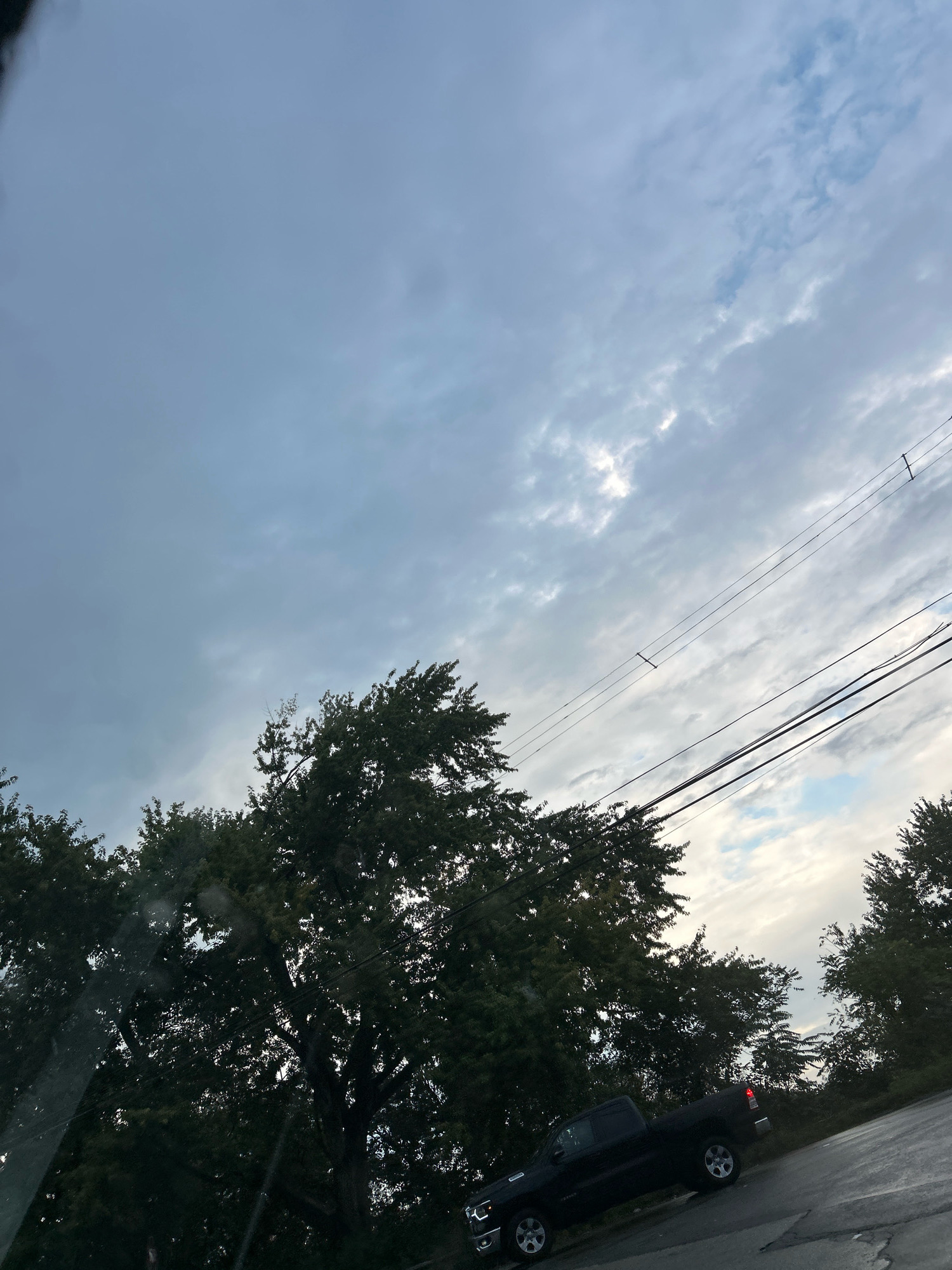 A black pickup truck is parked on a road in Robinson Township, USA. The truck is facing away from the camera and is positioned on the right side of the road. The sky above is cloudy with patches of blue sky visible. There are several trees on the left and right sides of the road, with their branches extending towards the sky. Overhead, there are power lines running across the scene. The road appears to be asphalt with some cracks visible.