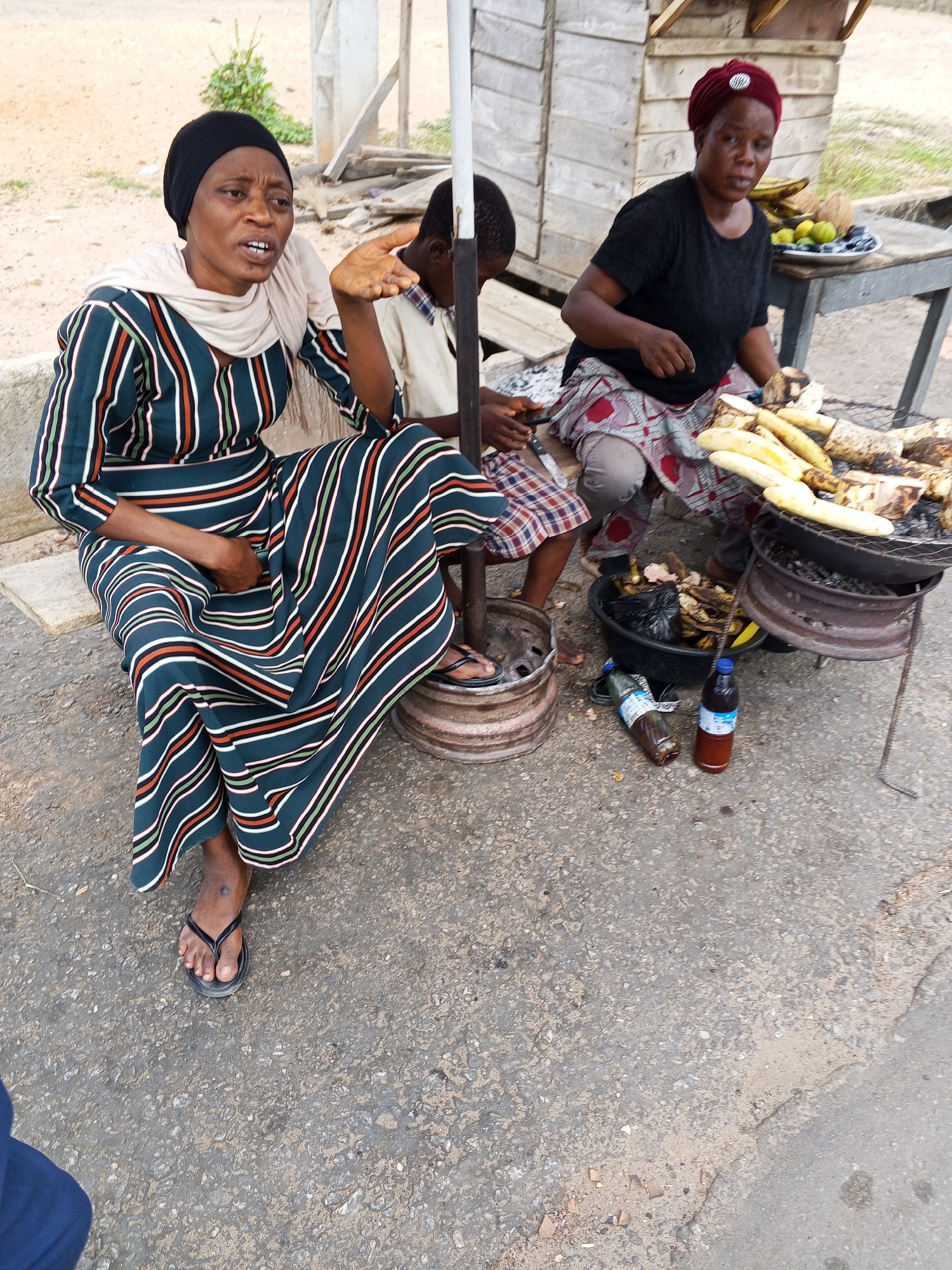 In Zango-ndaji, Nigeria, two women are present at a street-side food stall. One woman is seated, wearing a striped dress and a black head covering, engaged in a conversation with an animated hand gesture. Another woman wearing a black shirt, a red head covering, and a patterned skirt sits near a grill containing roasting plantains. There's a young person seated between them. The setting includes a black basin containing discarded plantains, along with two bottles placed on the ground. In the background there are buildings of wood and metal. The surface is concrete.