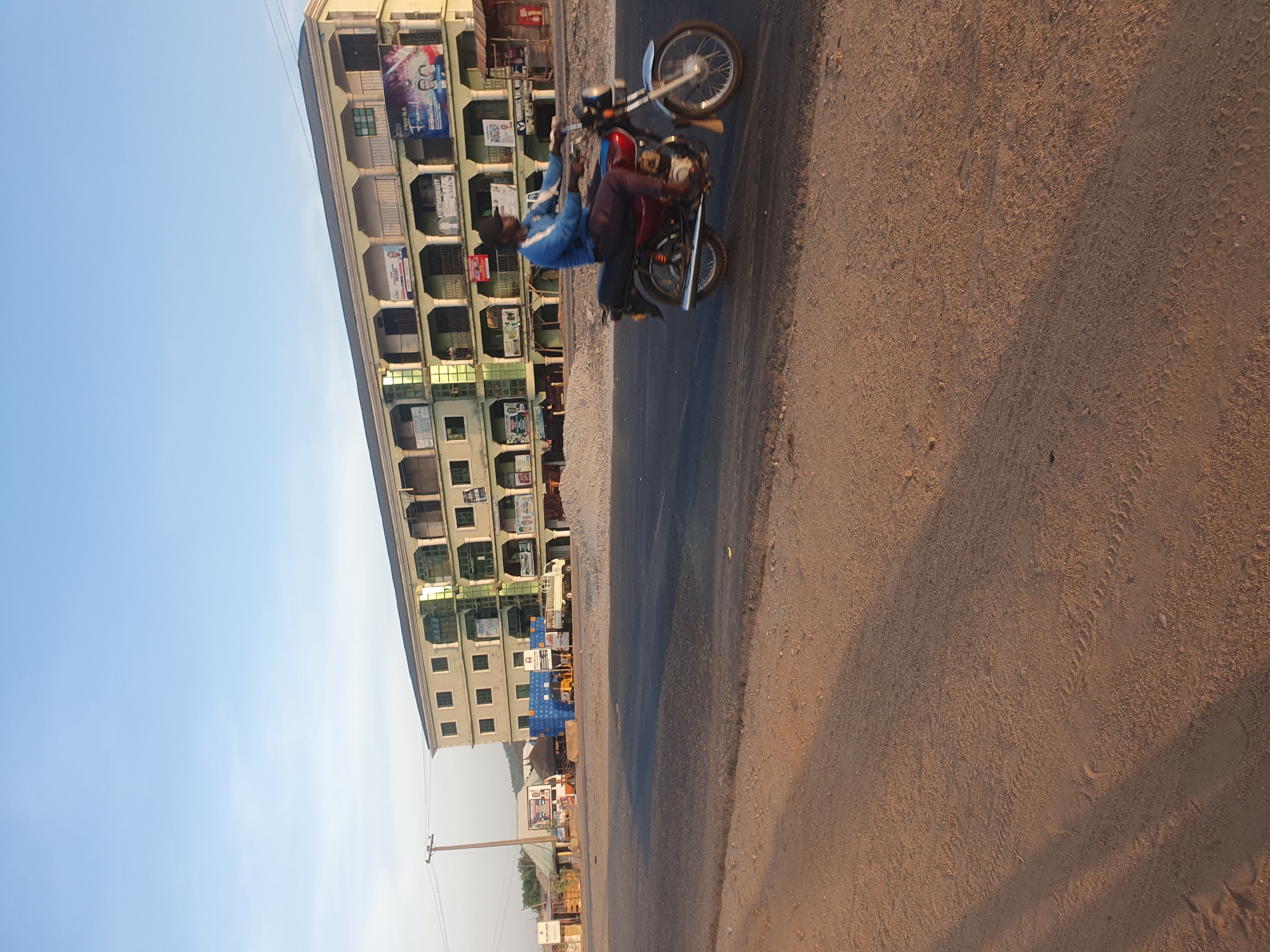 The image shows a motorcyclist riding on a road in Lokoja, Nigeria. The rider is wearing a blue shirt and dark pants. The motorcycle is red. In the background, there is a tall building with multiple balconies, each adorned with posters. The sky is clear and blue. The road appears to be asphalt with a dirt shoulder.