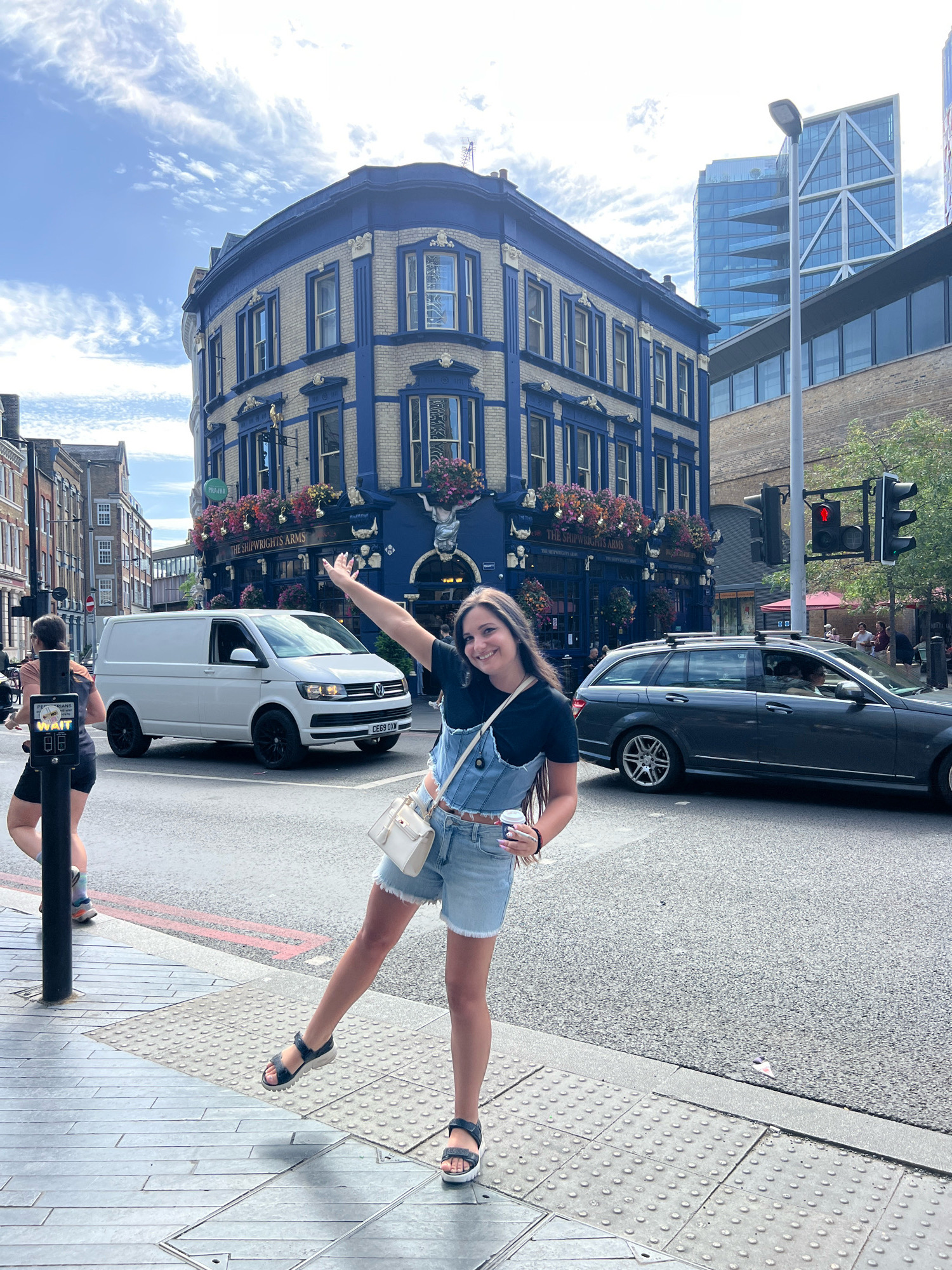 A young woman is posing for a photo in front of "The Shipwrights Arms" pub in London. She is wearing a denim overall dress and a black t-shirt, with a white purse slung over her shoulder and a cup of coffee in her hand. She is standing on a sidewalk with tactile paving, and a pedestrian crossing signal is visible in the background. A white van and a dark-colored station wagon are driving on the street. The pub is a prominent building with blue and beige facade, adorned with flowers. Modern glass buildings can be seen in the distance, indicating an urban environment. The sky is partly cloudy.