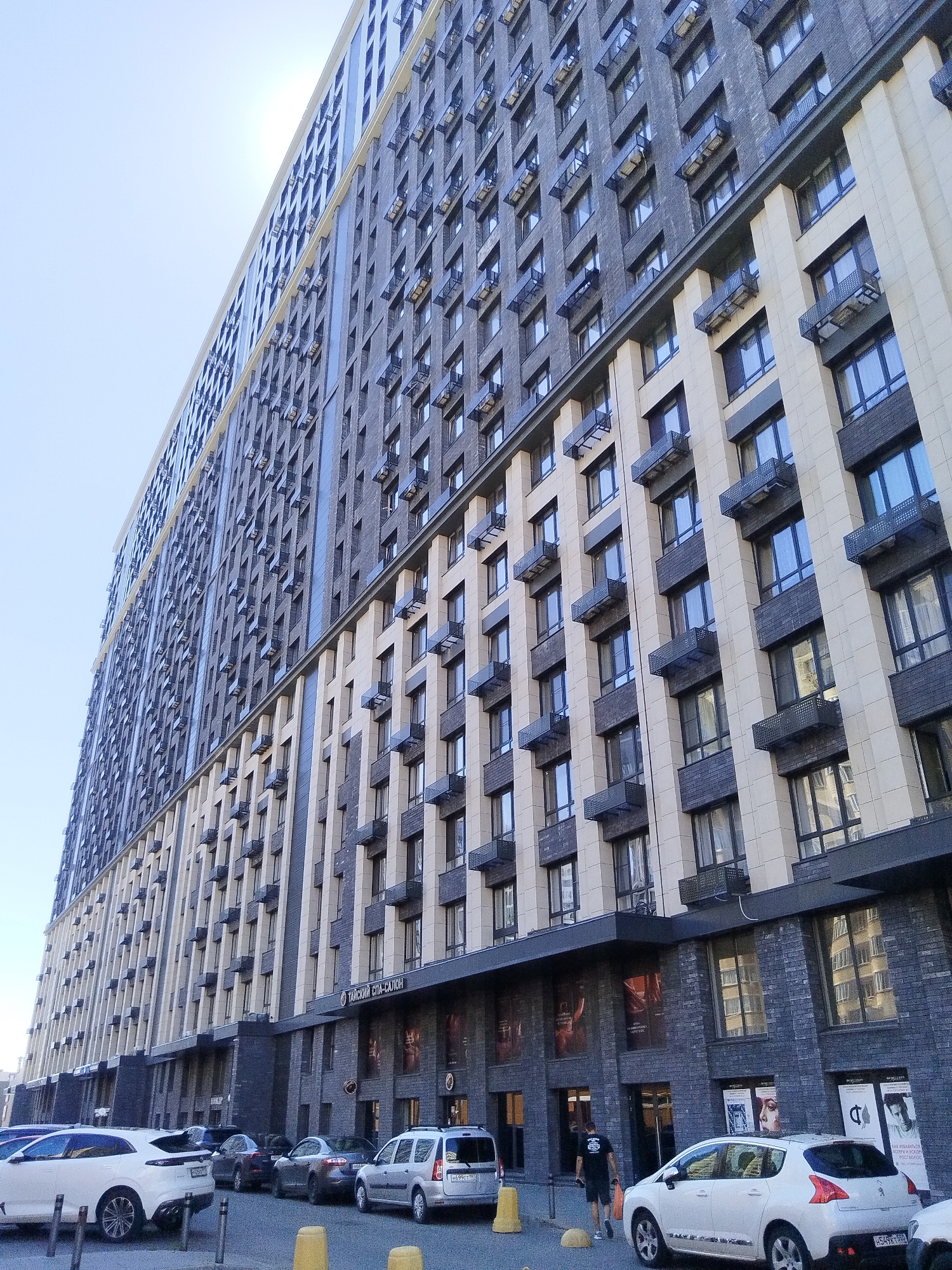 The image shows a modern, multi-story building with a facade of dark grey brick and light beige panels. Numerous windows, many with small black balconies or air conditioning units, are visible on the upper floors. The lower floors of the building appear to house commercial establishments, with signage visible, including what appears to be a sign in Russian that translates to "Thai Spa Salon". Several cars are parked along the street in front of the building, including a white SUV, a dark sedan, and a white hatchback. The sky is clear and bright. The scene is an urban environment in Mytishchi, Russia.