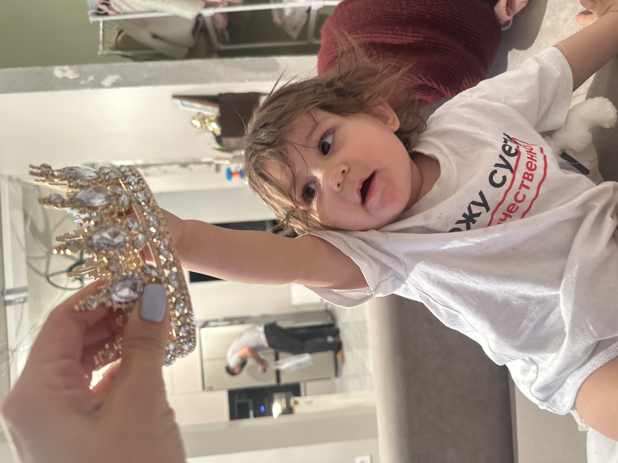 The image, rotated 90 degrees clockwise, depicts a tender indoor moment, likely in Moscow, Russia. In the foreground, an adult hand, adorned with light grey nail polish, extends forward, holding a shimmering gold-colored crown. This ornate crown is lavishly decorated with numerous clear, sparkling jewels.
The focus of the interaction is a young child, possibly a toddler, with expressive brown eyes and light brown hair, who is lying on their side. The child wears a white short-sleeved t-shirt, featuring prominent black Cyrillic text with a red outline. The text clearly reads "Я ЖИВУ ЕСТЕСТВЕННО," which translates to "I live naturally." The child's mouth is slightly open, and their gaze is fixed on the crown with an expression of intense curiosity and mild surprise. They are resting on a light-colored sofa or similar upholstered surface, with a dark red, textured fabric serving as a backdrop behind their head. A small, white, fluffy object is partially visible beside the child.
In the background, a blurred reflection in a mirror or a highly reflective surface shows an adult male, dressed in a light t-shirt and dark trousers, bent over. This reflection suggests an interior space with modern design elements, possibly a kitchen or an open-plan living area. The lighting indicates it is daytime indoors. The overall atmosphere conveyed is one of playful interaction and affection.