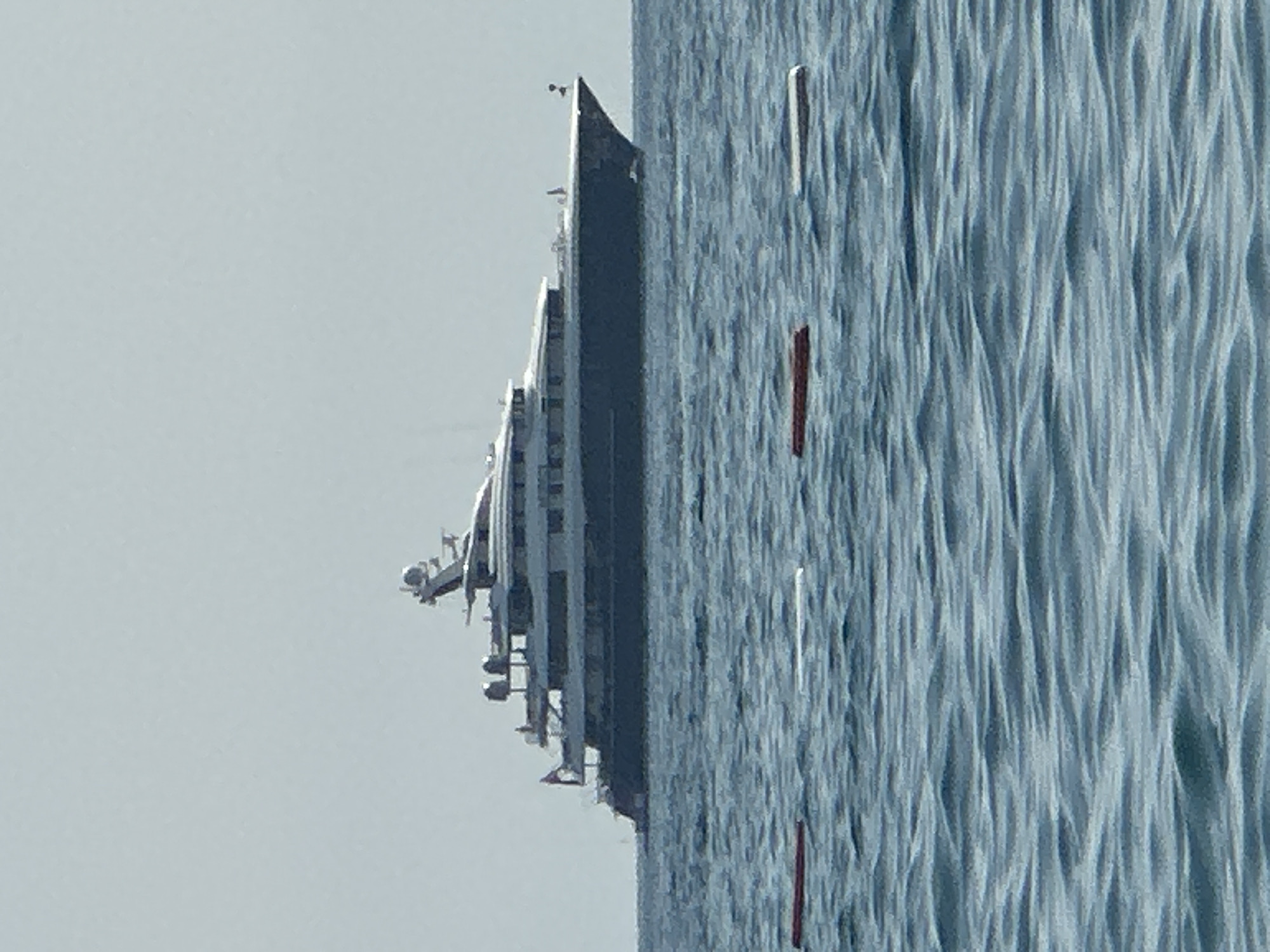 A large, dark-hulled yacht with multiple decks and a white superstructure is anchored in calm, blue water. The yacht has a sleek, modern design, with a visible radar dome and antenna on its upper deck. In the foreground, there are several floating markers, some red and some white, on the surface of the water, indicating a swimming or boating area. The sky is a pale, hazy gray. The scene appears to be a peaceful day at sea, likely off the coast of Boreti, Montenegro, known for its luxury yachts and coastal beauty. No people are clearly visible on the yacht or in the water. The overall impression is one of opulence and tranquility.