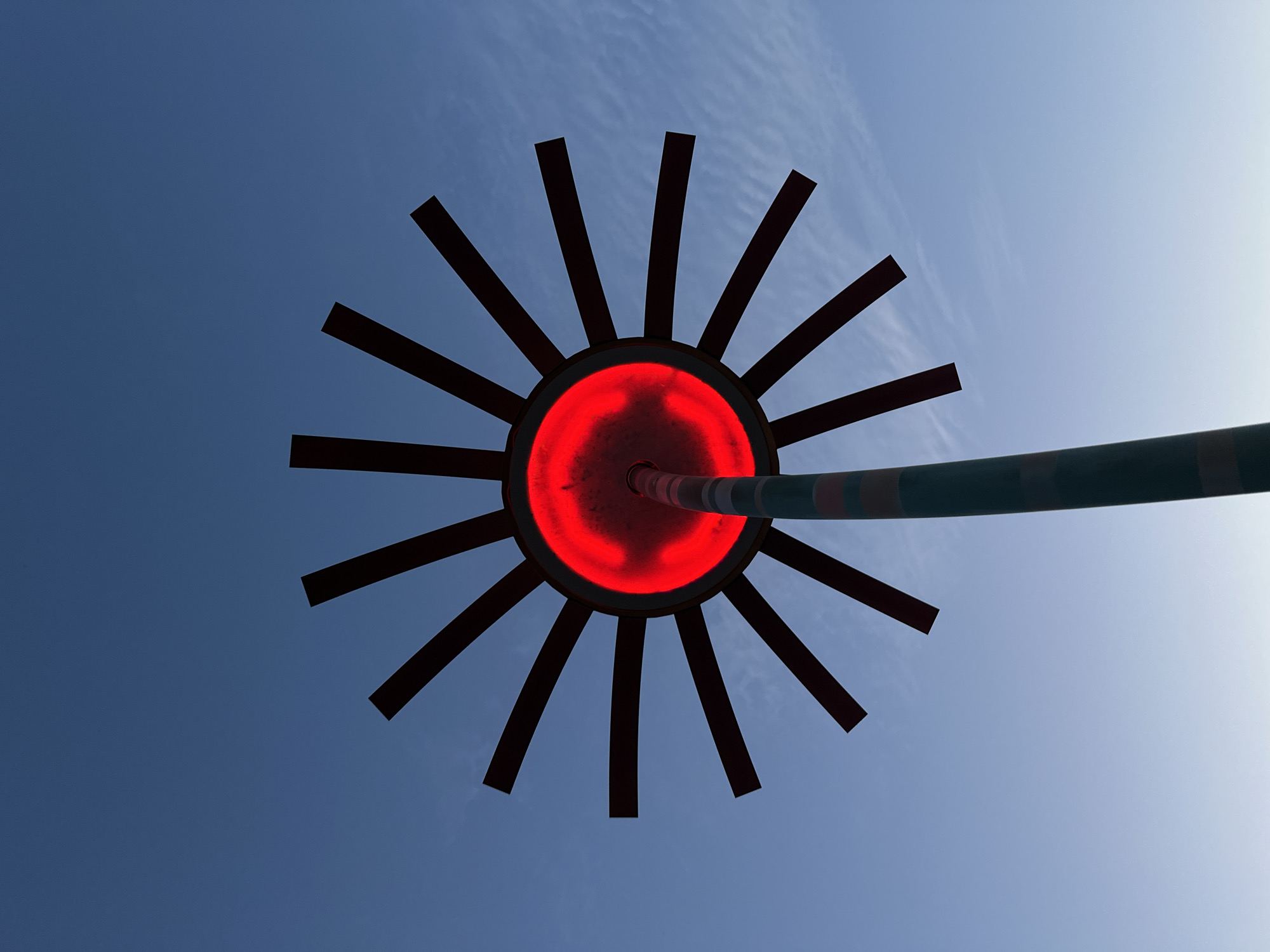 The media file shows a low-angle shot of a circular object with radiating spokes against a bright blue sky with wispy clouds, suggesting daytime. The central part of the object emits a vibrant red light, and a dark pole extends from its side, banded with green and red stripes. The object appears to be an illuminated sculpture or part of a structure, possibly a stylized representation of the sun or a decorative lamp. The sky is clear and bright, indicating fair weather. The overall impression is abstract and graphic, with strong contrasts between the dark spokes, the glowing red center, and the expansive sky. No people are visible, and the setting is undefined beyond the open sky. The time of day is clearly daytime due to the bright sky, and the clouds suggest it might be a slightly hazy or humid day. The image's perspective, looking upwards, emphasizes the scale and design of the object against the vastness of the sky.