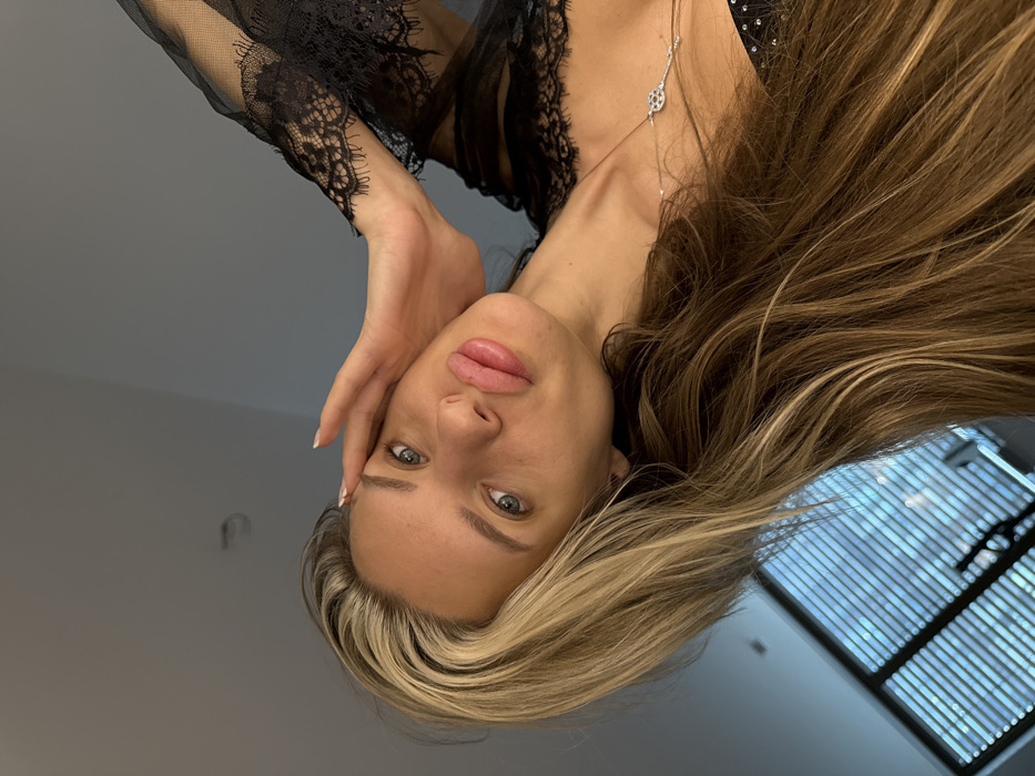 The image displays a close-up, tilted portrait of a female subject indoors. The subject has long, multi-toned blonde and brown hair, blue eyes, and full, slightly parted lips. Her right hand is raised, with fingers resting on her temple; her fingernails are light-colored. She wears a black garment featuring lace on the left sleeve, possibly a robe or lingerie, and a black top or bra with small, reflective studs. A delicate necklace with a circular, open-work silver-toned pendant is visible on her chest. The subject is looking directly at the camera. The background consists of a plain light grey ceiling in the upper left, a darker plain grey wall on the right, and a window with horizontal blinds in the lower left, through which diffused outdoor light is faintly discernible. The specific location within Berlin, Germany, cannot be determined from the visual information provided.